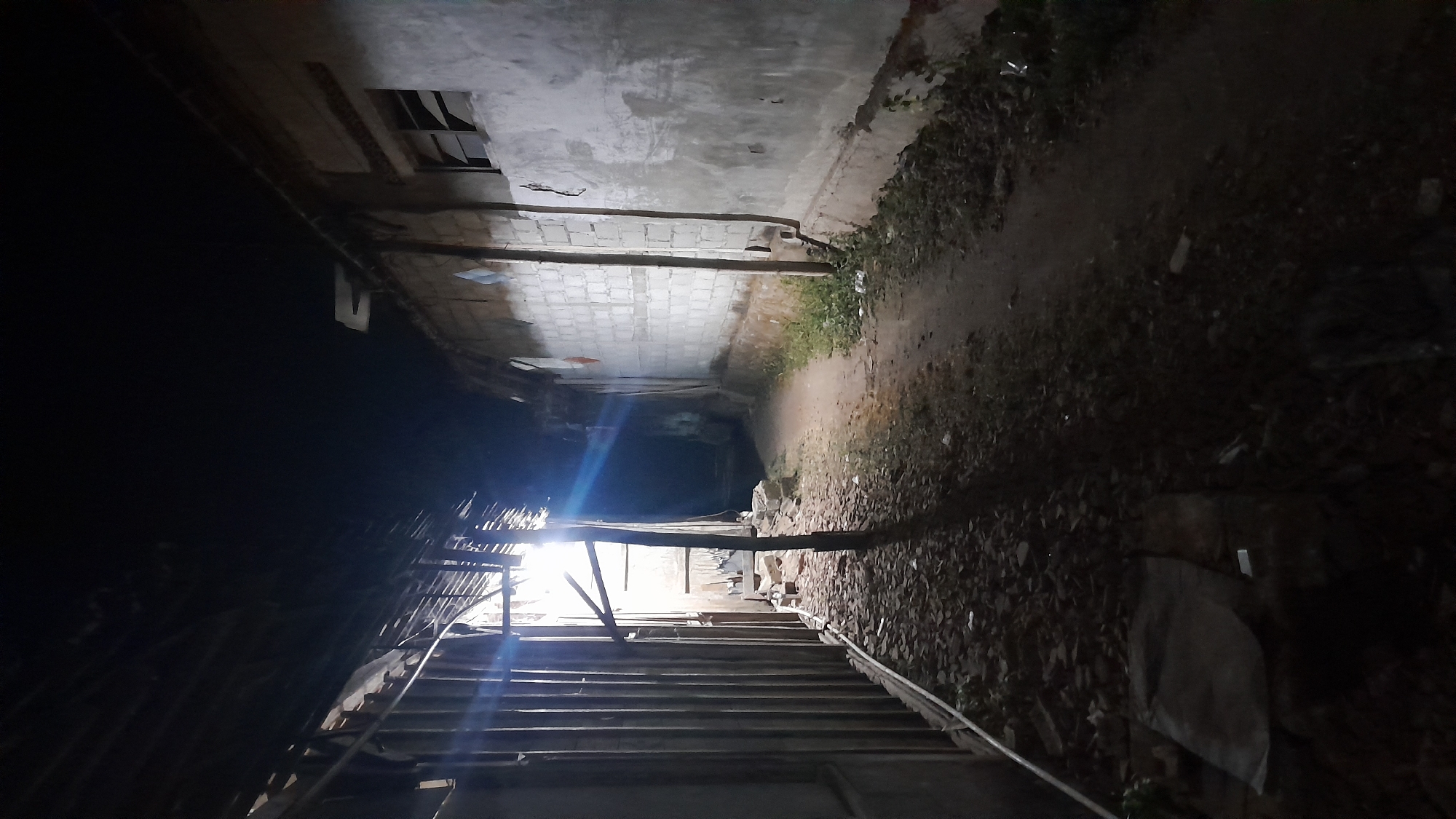 The image depicts a narrow, unpaved alleyway at night in the city of Sumberarum, Indonesia. On the left, a dilapidated structure with a corrugated metal roof and vertical wooden planks lines the alley. Debris, including leaves and what appears to be broken building materials, is scattered on the ground. A bright light source, possibly from within the structure or a nearby lamp, casts strong shadows and creates a lens flare effect.
On the right side of the alley is a wall constructed of concrete blocks, partially plastered and stained. There is a broken window on this wall. The ground on this side is covered with some vegetation and dirt. The alley recedes into darkness in the distance. The overall scene suggests an impoverished or neglected urban environment. There are no people visible in the image.