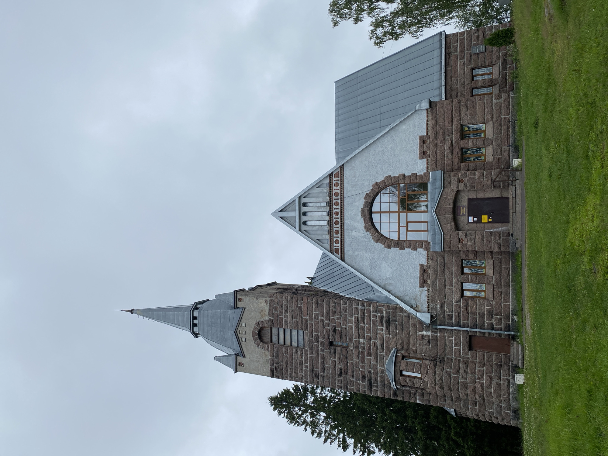 The image shows the exterior of a stone church in Melnikovo, Russia, under an overcast sky. The church has a prominent bell tower with a pointed grey roof and a spire. The main building features a large, arched window with a wooden frame and multiple panes, situated above the entrance. The walls of the church are constructed from rough-hewn, reddish-brown stones, with lighter grey stucco sections on the upper parts of the main building and the bell tower. Several smaller windows with stained glass are visible on the lower levels. The church is surrounded by green grass, with trees visible to the left and right. The overall impression is of a historic, solid structure set in a natural, somewhat rustic environment.