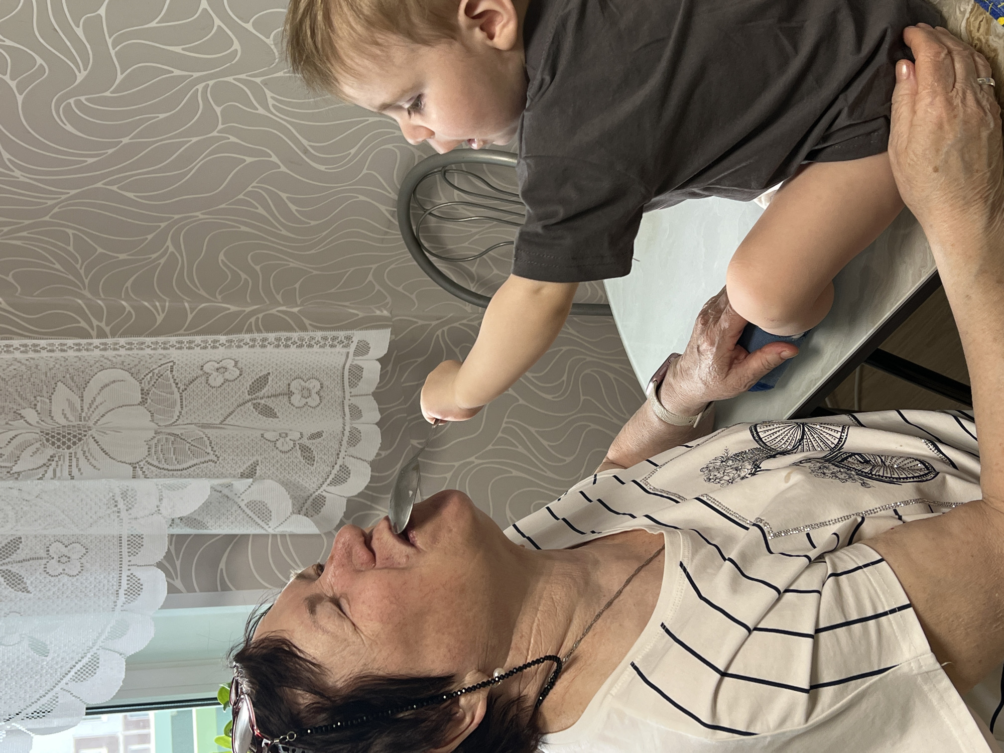 This image captures a candid moment between a young child and an older woman indoors, in what appears to be a home setting (context provided by the user as Perm, Russia).
The main subjects are a toddler-aged child and an adult woman. The child, with light hair and wearing a dark t-shirt, is standing on a light-colored table. The child looks focused downward, possibly reaching for or playing with something on the table surface. The woman, with dark hair, glasses, and wearing a striped top, is supporting the child by holding their legs/lower body. She is looking up at the child with her mouth open, suggesting she might be speaking, laughing, or singing to the child. Her hands, showing age, are gently holding the child, indicating a caring interaction.
The background features a light-colored wall with a repeating, wavy, organic pattern. To the left, white lace curtains with floral patterns are visible, suggesting the presence of a window. A metallic chair back is partially seen behind the child. The overall atmosphere conveys warmth and familial affection. No text is visible in the image.