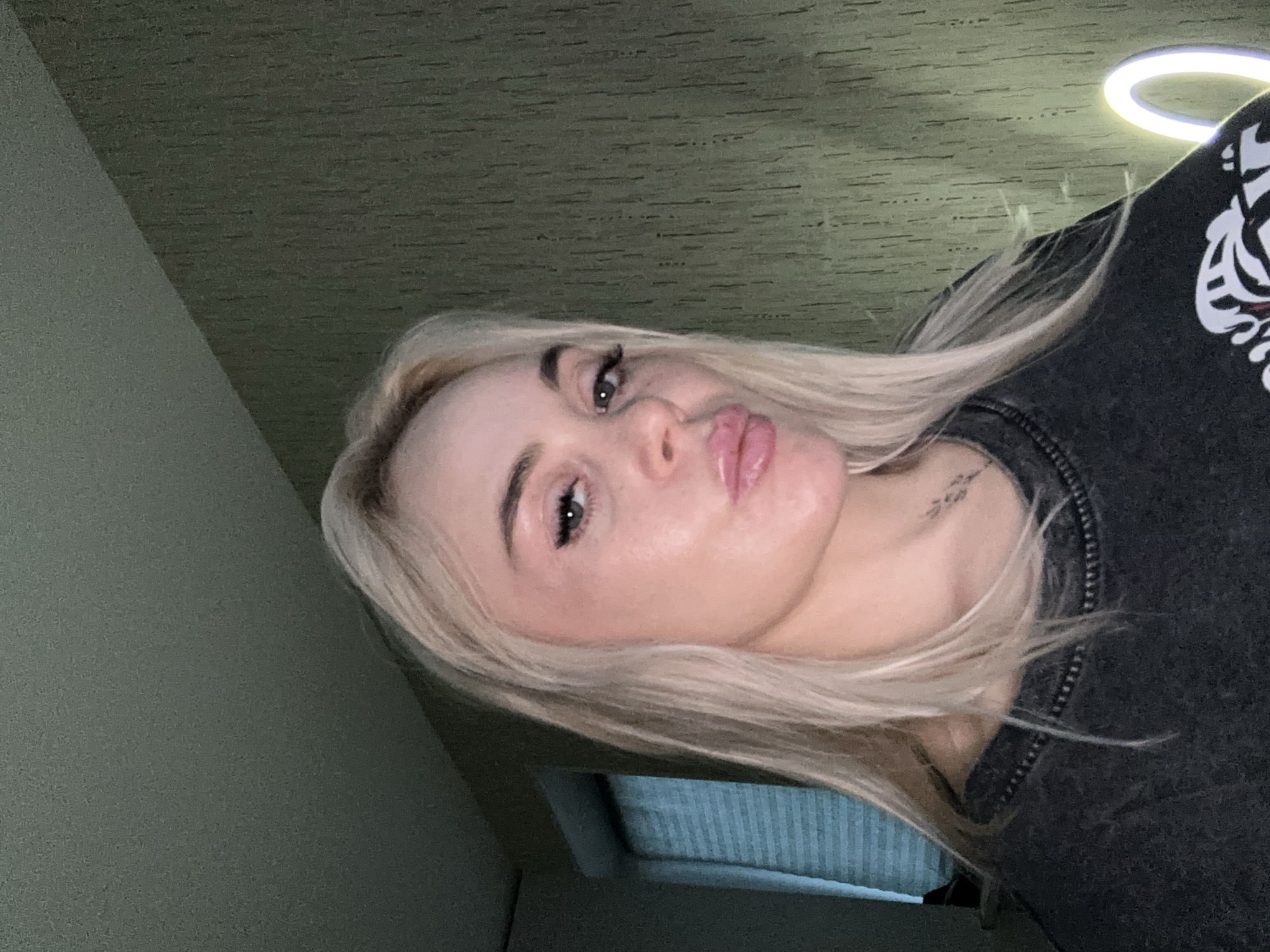 A close-up selfie captures a person with long, flowing blonde hair, angled sideways in the frame. The subject, likely female, is looking towards the viewer with her head tilted, making a "puckered lips" expression. She has noticeable eye makeup, including winged eyeliner and mascara, and her lips appear glossy. A small, indistinct tattoo can be seen on her neck, just below her right ear. She is wearing a dark-colored top, possibly a sweatshirt, which features a white graphic design on the right shoulder, resembling part of a tiger or a similar animal.
The background consists of a textured, dark olive green surface, appearing to be a ceiling. In the upper right corner, a bright, circular light source, consistent with a ring light, is visible. The lower left portion of the image shows a hint of another interior space, possibly a window with vertical blinds or a wall with a textured pattern. The overall lighting suggests an indoor setting. The image is oriented vertically but the subject's head is rotated approximately 90 degrees clockwise. No discernible text is visible beyond the faint tattoo.