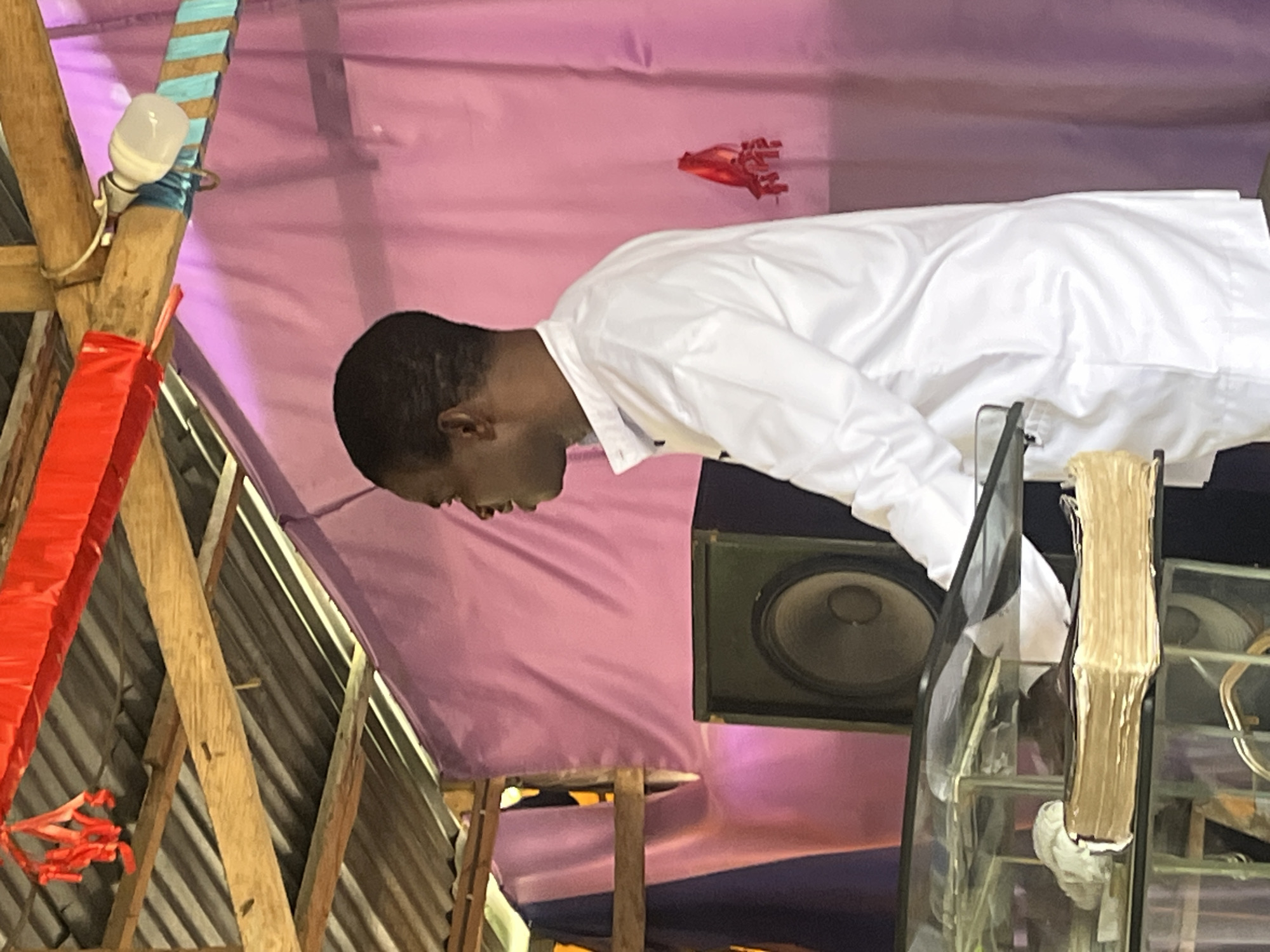 A man in a white shirt is standing at a pulpit in Iranla, Nigeria. The pulpit is made of glass. A speaker sits on the side of the pulpit. The background is a pink tarp. The ceiling is corrugated metal. A string of red flags hangs from the ceiling.