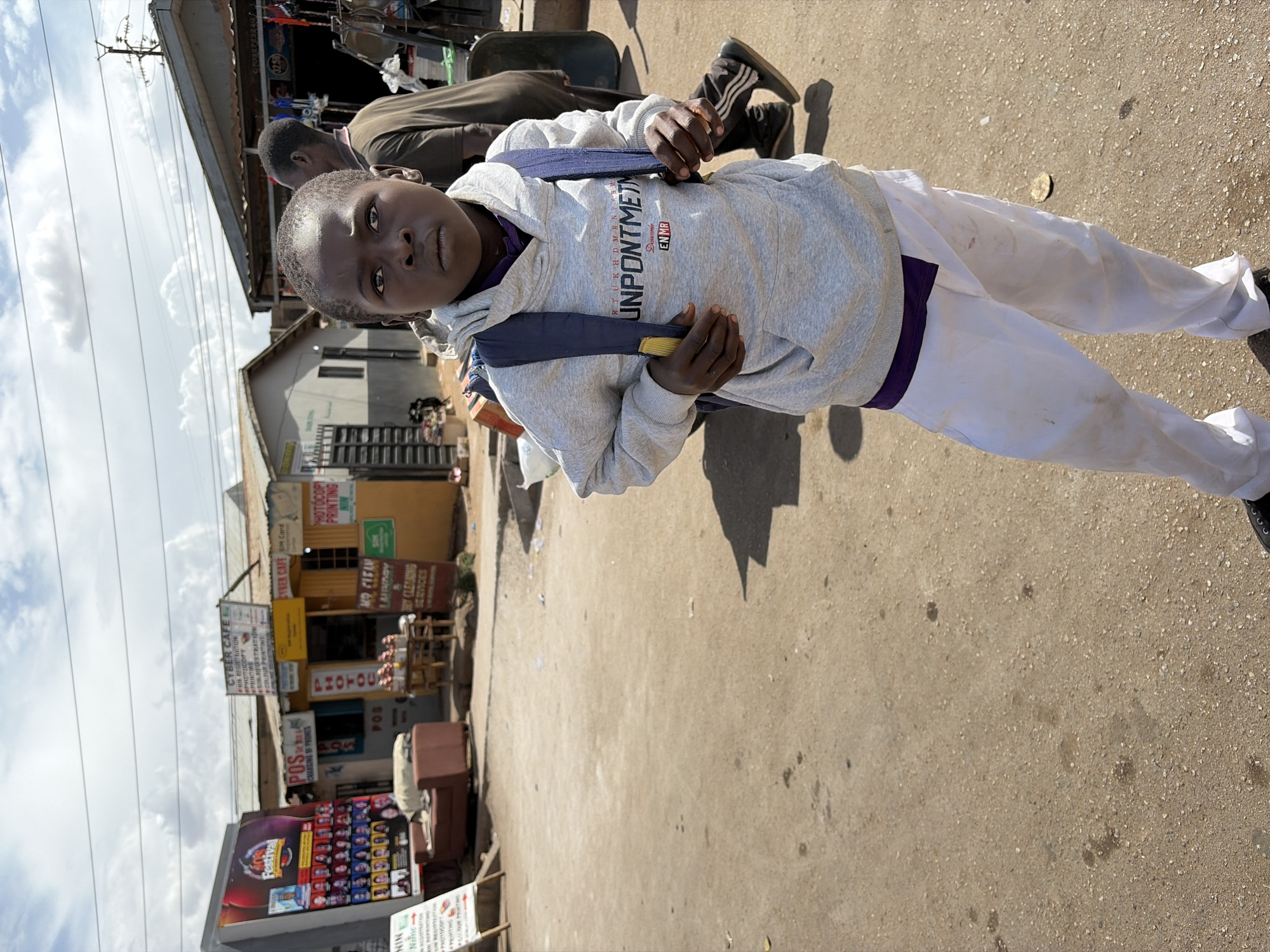 A portrait-style shot captures a young person standing on a paved street in Jos, Nigeria, looking directly at the camera. The subject has dark skin and short, closely cropped hair, wearing a grey hooded sweatshirt with a blue backpack, white pants, and dark shoes. The hoodie features text that appears to read "UNPONTMETM" and "EN MR". Their expression is neutral, and their hands are on the straps of the backpack.
In the background, an adult person is partially visible, walking away from the camera. The setting is a bustling street lined with modest, single-story commercial buildings and stalls. The ground is a light-colored asphalt or concrete, showing some wear and scattered debris. Above, the sky is bright with scattered clouds, indicating a clear day.
Numerous signs are visible on the buildings, advertising various services. These include "PHOTOCOPY PRINTING," "CYBER CAFE" offering services like "SIM Card" and "NIN REGISTRATION," "POS CARD LOADING OF PHONES," and "DRY CLEANING SERVICES." One sign also mentions "Festival." The overall scene depicts a typical commercial street activity during daytime.