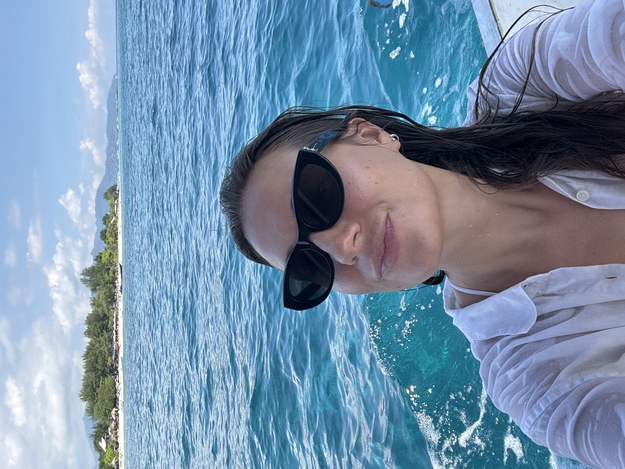A woman wearing sunglasses and a white shirt is on a boat, with the turquoise ocean and a beach in the background. The location is possibly in the city of Unknown, Indonesia, based on the image's title and file name. The woman's hair is wet, and water splashes near the boat. The beach has trees, sand, and possibly buildings.