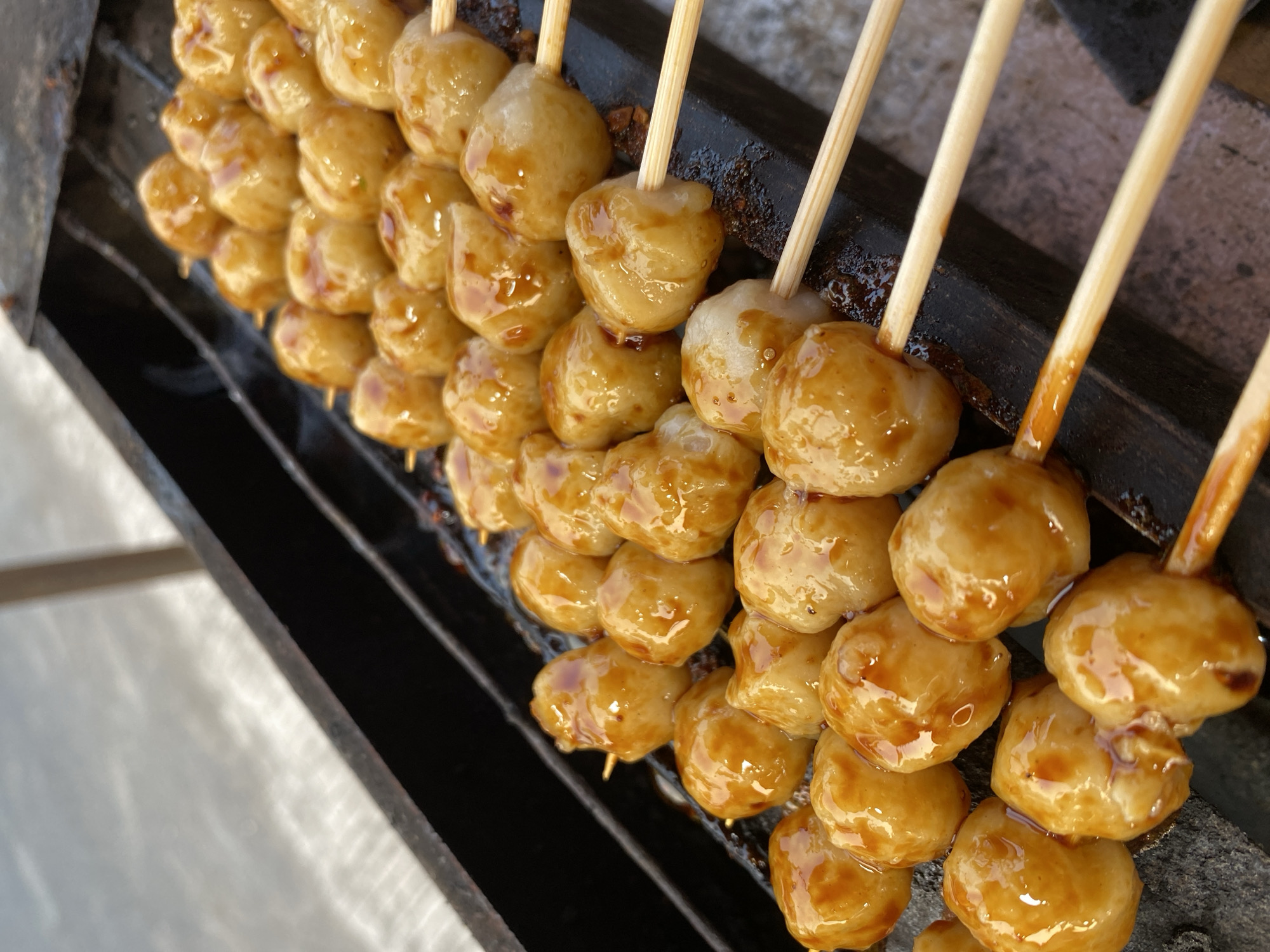 The image shows several skewers of meatballs being grilled over a dark surface, likely a grill. The meatballs are small, round, and coated in a glossy, dark sauce. They are arranged in rows on wooden skewers. The background is blurred, but appears to be an outdoor setting. The overall impression is one of food being prepared for sale or consumption, possibly at a street food stall.