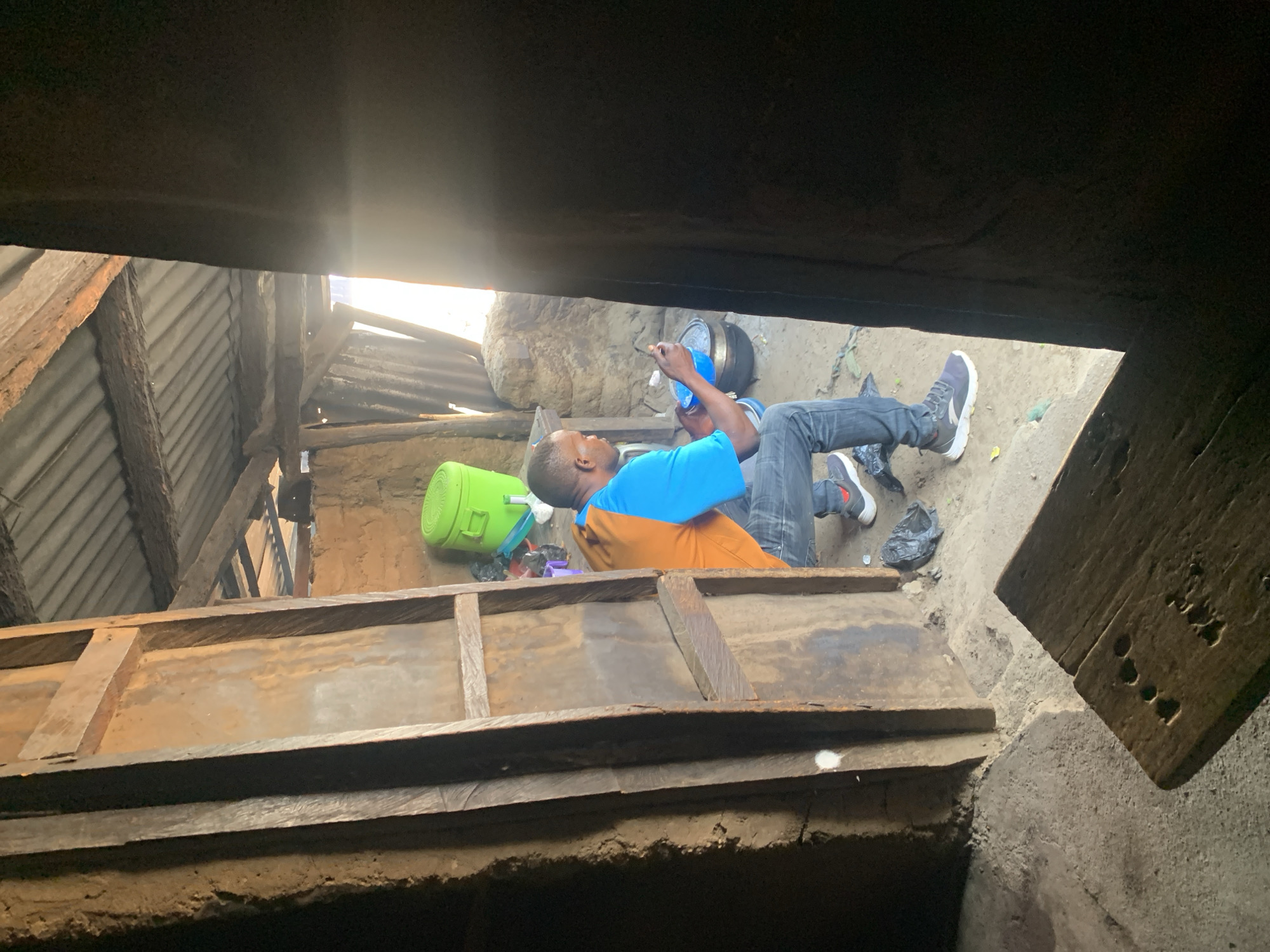 In a dimly lit, rustic interior of what appears to be a dwelling in Project, Nigeria, a man wearing a blue and orange shirt and denim jeans is squatting. He is holding a blue bowl and a utensil, seemingly engaged in an activity involving a pot situated on the ground. To his left, a bright green bucket is visible. The environment is characterized by mud walls, a corrugated metal roof supported by wooden beams, and a dirt floor. The scene suggests a simple living condition, possibly a kitchen or a common area within a home.