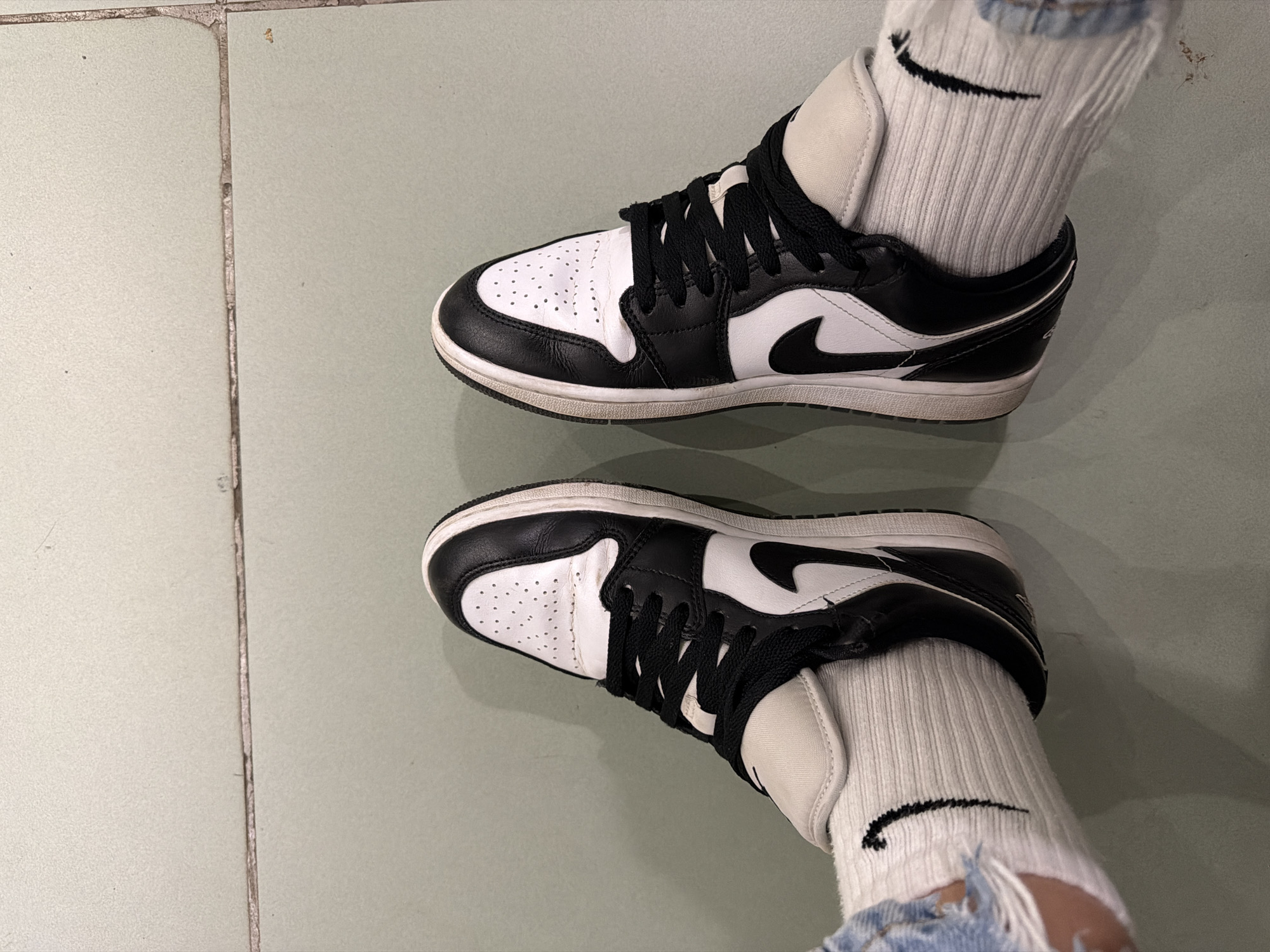 A top-down shot shows a pair of feet wearing black and white Nike Air Jordan 1 Low sneakers and white Nike logo socks. The person is wearing distressed light-wash denim jeans. The location appears to be indoors on light green tile flooring, possibly in Sofia, Bulgaria, but the exact location cannot be determined.