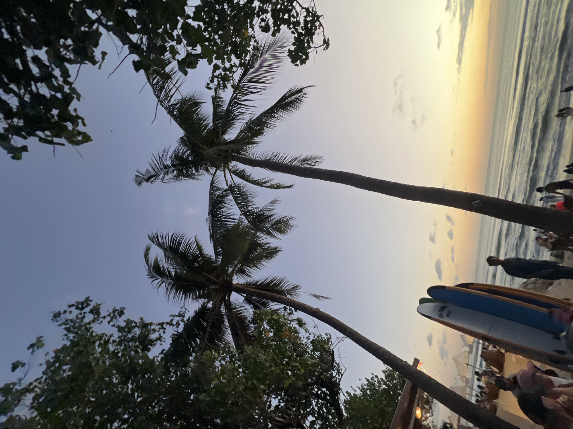 The image depicts a tropical beach scene in Kuta, Indonesia, likely at sunset or sunrise. Two tall palm trees dominate the foreground, their fronds silhouetted against a gradient sky that transitions from a deep blue at the top to soft orange and yellow hues near the horizon. In the middle ground, several surfboards stand upright on the sandy beach, leaning against each other. To the left, a beachfront establishment with a thatched roof and illuminated lights is visible, with people seated at tables, enjoying the view or a meal. A few individuals are scattered on the beach, some walking and others standing near the surfboards. The ocean stretches out to the horizon, with gentle waves rolling onto the shore. The overall atmosphere is relaxed and idyllic, characteristic of a popular tourist destination.