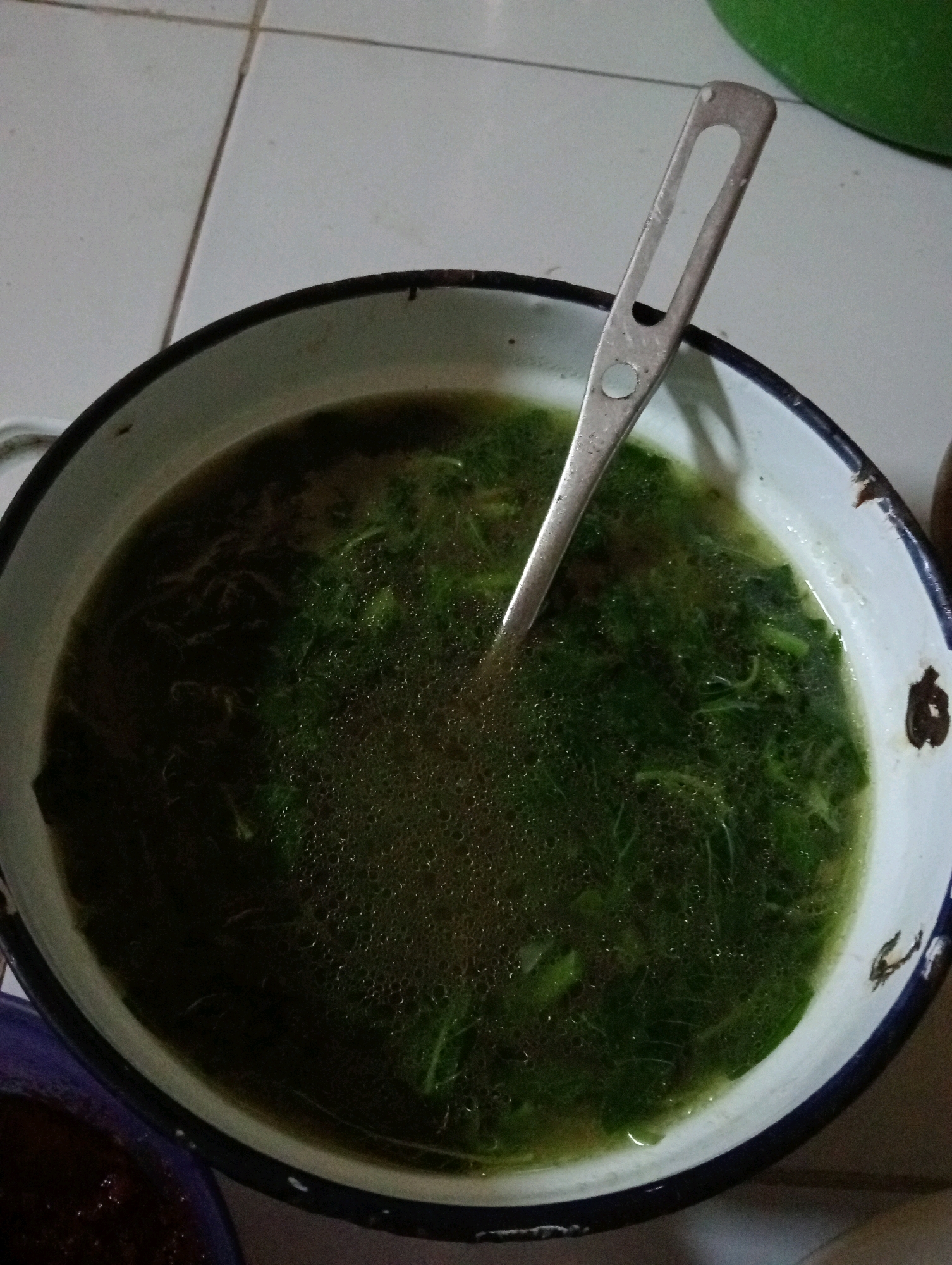 A metal bowl filled with green soup and vegetables sits on a tiled floor. A metal spoon is placed in the soup. A portion of a purple bowl with red food is visible on the left side of the image, and a green object is visible on the right. The setting appears to be a domestic environment in Pagerungan Kecil, Indonesia.
