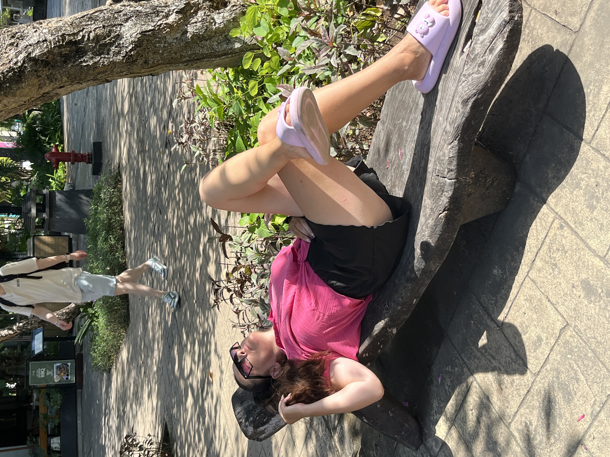 In Kuta Selatan, Indonesia, a woman with dark hair and sunglasses lies on a rustic wooden bench, her head tilted back and eyes closed, appearing to be sunbathing. She wears a bright pink short-sleeved shirt and black shorts. Her left leg is bent at the knee, and she is wearing a light purple slide sandal with a decorative detail on the strap. A second person, a man wearing a light-colored t-shirt, light blue shorts, a backpack, and athletic shoes, walks away in the background. The environment is a paved outdoor area with dappled sunlight filtering through trees, creating shadows on the ground. Various plants and greenery surround the scene, and a red fire hydrant is visible in the middle distance.