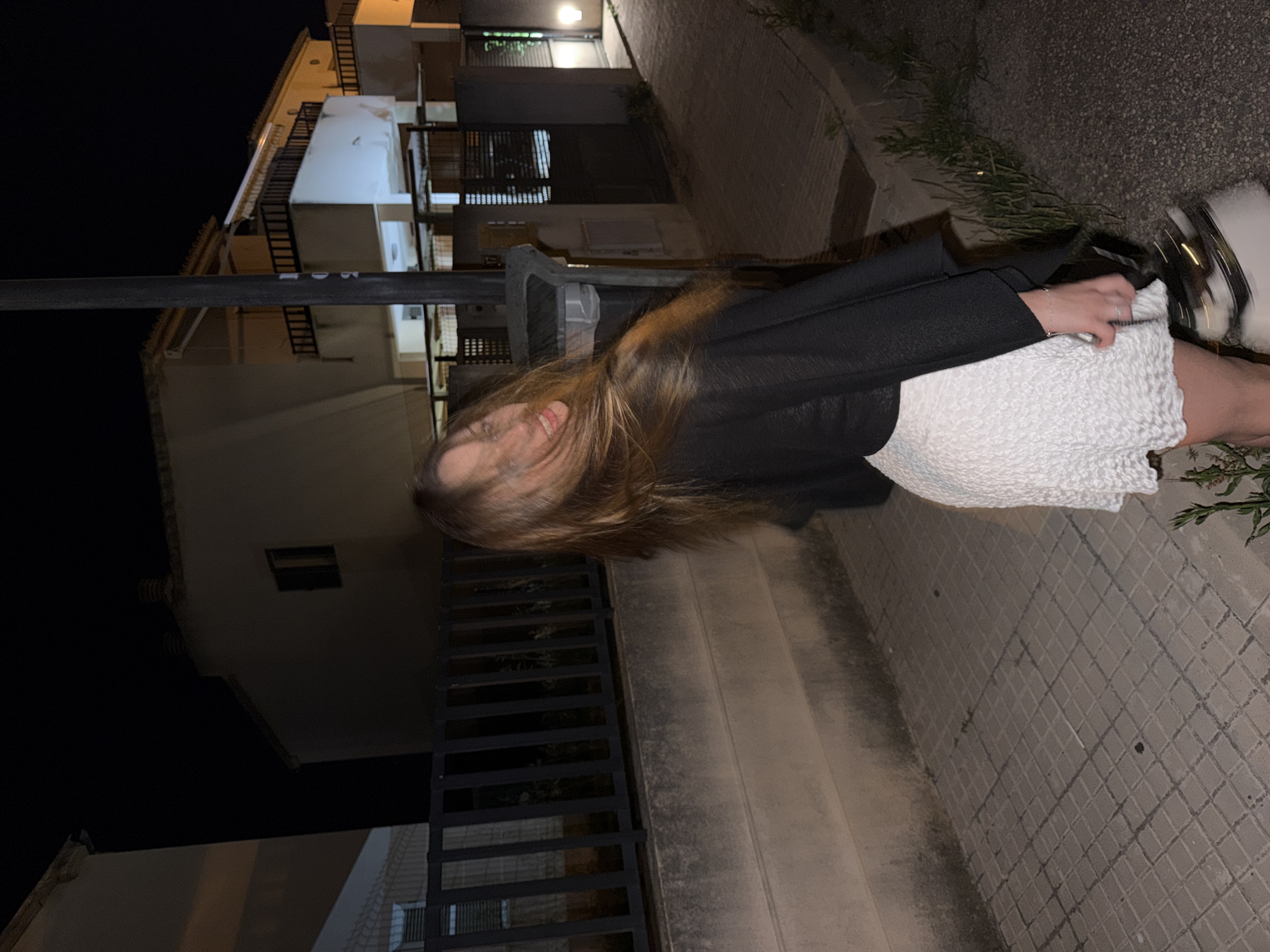 A young woman with brown hair is standing on a sidewalk next to a building at night in Platja D'alcúdia, Spain. She is wearing a black jacket and a white textured dress, and white sneakers. She is looking toward the camera and smiling slightly. The sidewalk is made of gray brick. There are green plants growing along the edge of the sidewalk. Buildings are visible in the background.