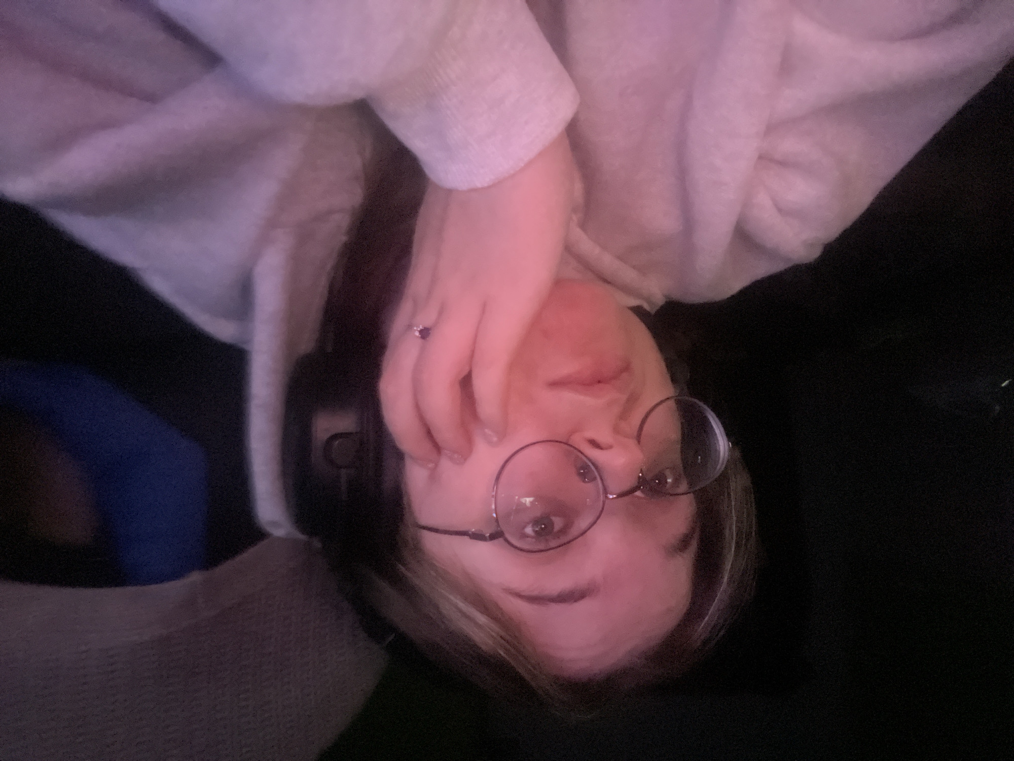 A young person wearing round eyeglasses and a light-colored hooded sweatshirt is looking towards the camera. Their left hand is resting on their cheek, and a ring with a dark-colored stone is visible on their ring finger. The lighting in the scene is dim with a pinkish hue. The background is dark and indistinct, with hints of a textured surface, possibly a chair or a headrest, on the right side. The image does not provide any information or context to suggest a location in the city of Kolomna, Russia.