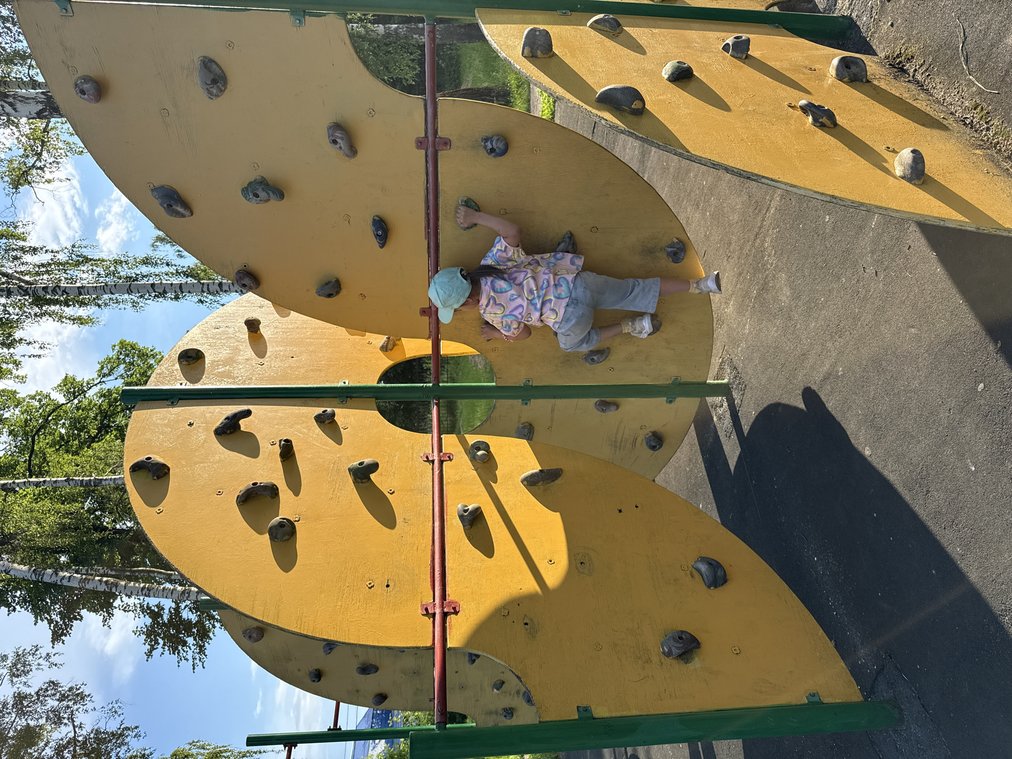 A young girl is climbing a yellow artificial rock-climbing wall on a sunny day. She is wearing a light blue cap, a colorful t-shirt with hearts, and light blue jeans. The wall is a curved structure with various climbing holds. The background shows trees and a bright blue sky with some clouds. The girl is positioned mid-climb, with her hands and feet on the holds. The scene appears to be in a park or playground.