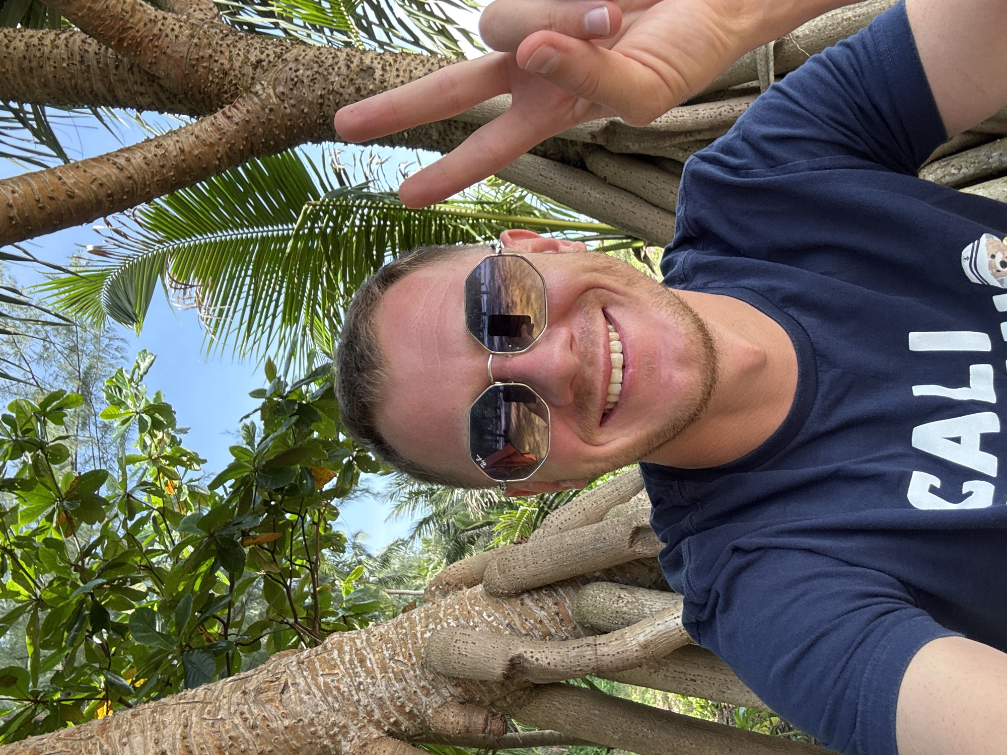 A man with short blond hair, wearing sunglasses and a navy blue t-shirt, smiles and flashes a peace sign with his right hand. He is positioned in the foreground, with large tree roots and lush green foliage forming the background. The man's sunglasses reflect a scene of trees and a blue sky. The overall setting appears to be a tropical outdoor environment, possibly near a beach, given the presence of palm fronds and the clear blue sky. The man's t-shirt has white lettering, partially visible, that reads "CALI" and a graphic of a teddy bear wearing a hat. The image seems to be taken in Ban Khuk Khak, Thailand, suggesting a vacation or leisure activity in a warm climate.