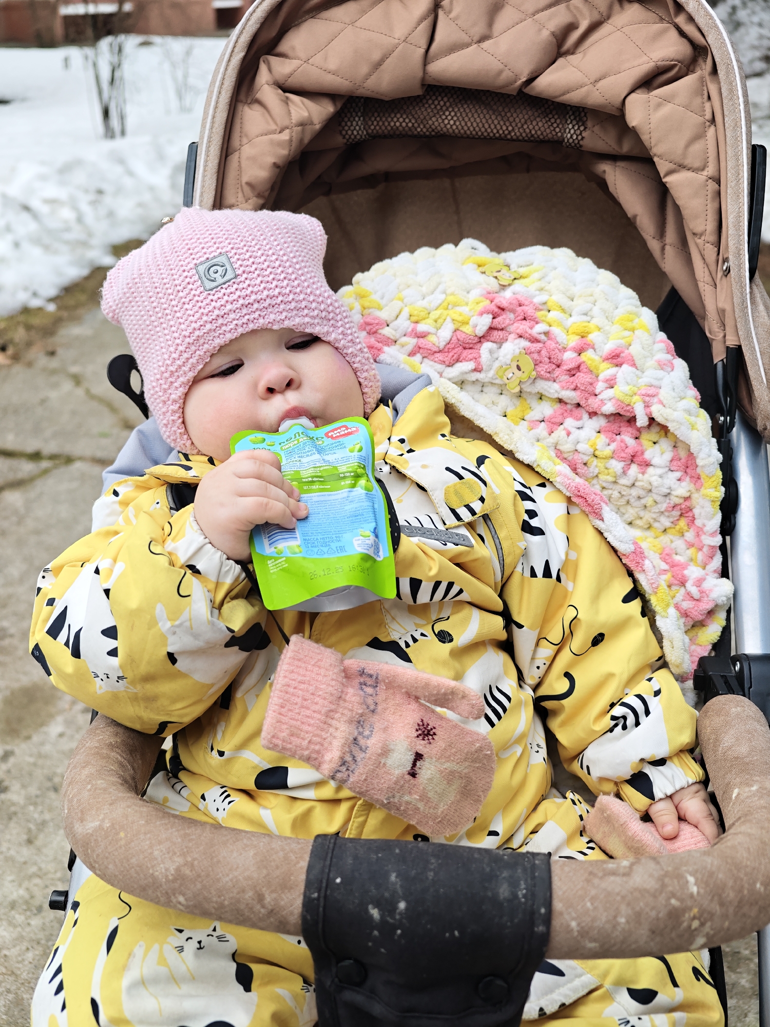 A baby sits in a stroller while drinking from a green and blue pouch of baby food. The baby wears a pink knit hat and a yellow snowsuit printed with white cats and black stripes. A pink and white knit blanket lies next to the baby, and a pair of pink mittens rests on the baby's lap. The stroller is beige and brown. Patches of snow are visible in the background, suggesting a cold climate. The baby food pouch features Cyrillic script and the date "26.12.25," indicating the food may be of Russian origin. Based on the provided OCR text, it appears to be from a product called "пюре да" (Puree Yes) from the city of Chaikovsky, Russia.