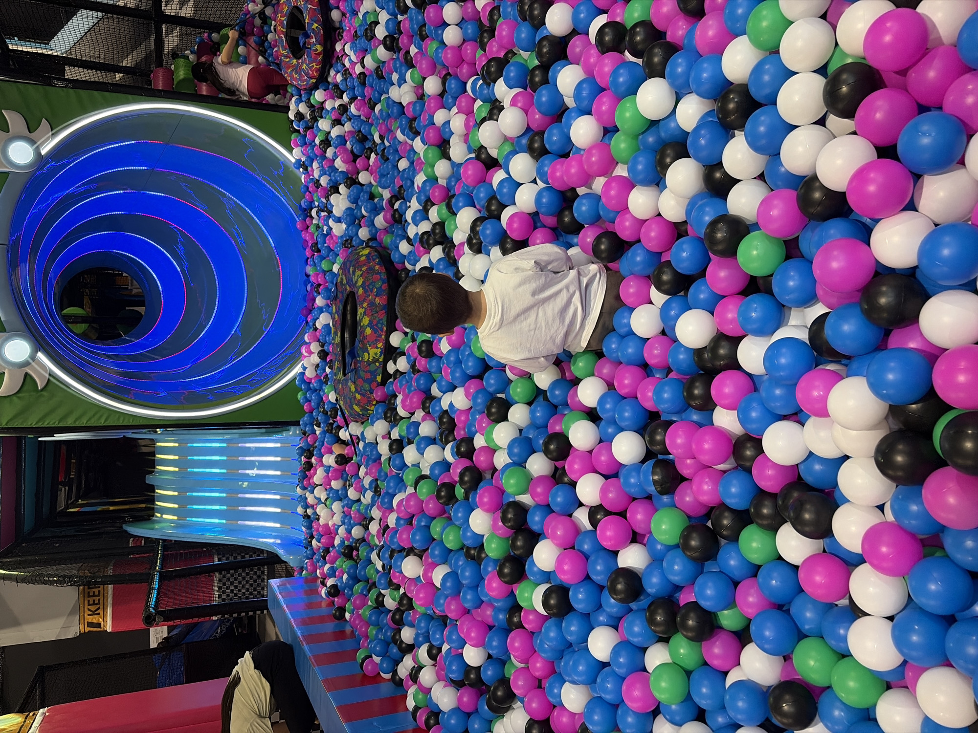 A ball pit with a boy wearing a white shirt sitting within it. The pit contains blue, pink, white, green, and black balls. Next to the pit is a slide and a blue-lit tube. In the top left corner, an individual is lying on a colorful inflatable tube. The location is likely an indoor playground. The city of Sofia, Bulgaria is not discernible from the image.