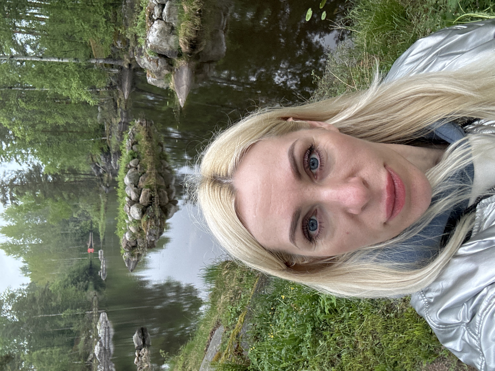 A medium shot captures a woman with blonde hair and blue eyes, who appears to be taking a selfie. She is wearing a silver jacket and is positioned in front of a body of water with rock formations and trees visible in the background. The scene is set in a natural environment, likely a park or garden. The location is the city of Unknown, Unknown.