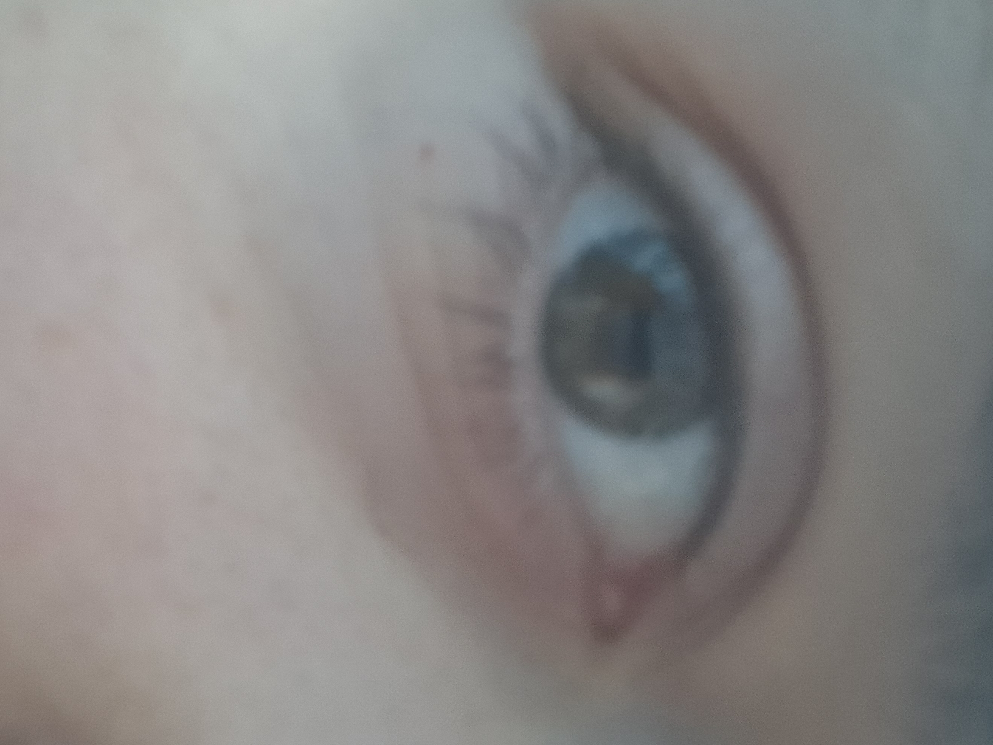 The image shows a close-up, out-of-focus view of a human eye. The iris appears to be a muted greenish-brown color. The surrounding skin is visible, with some subtle texture and a hint of redness below the eye. Eyelashes are present but blurred. The lighting is diffuse, creating soft shadows and a lack of sharp detail. The background is entirely out of focus and consists of indistinct, muted tones. There are no identifiable objects or features that would suggest a specific location, city, or environment.