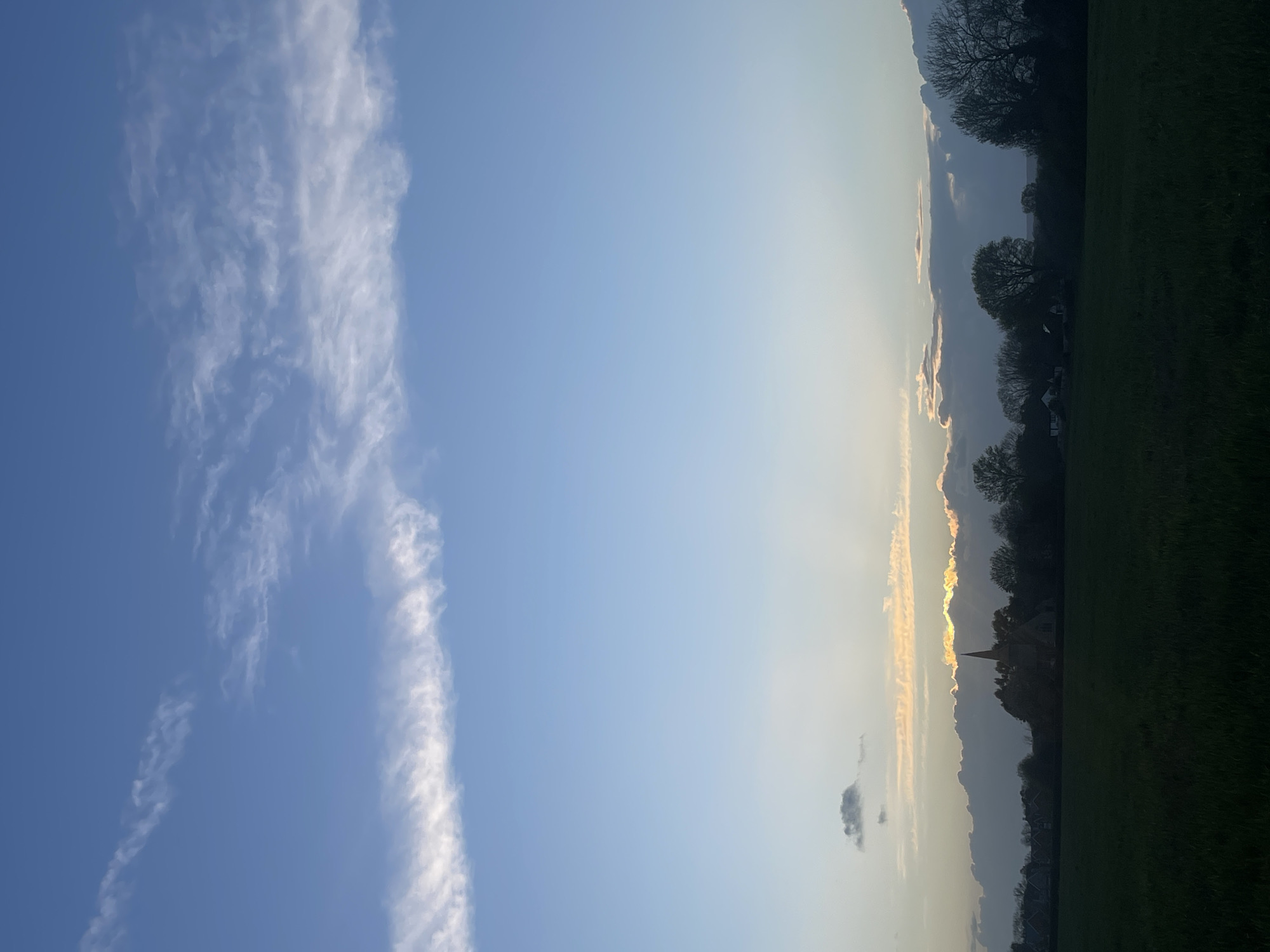 The image shows a landscape in Maidstone, United Kingdom. The sky, predominantly blue, is marked by elongated white clouds on the left. Toward the right, the sky transitions to a lighter hue, hinting at either dawn or dusk. A dark treeline stretches across the right side of the image, silhouetted against the sky. A church spire is visible in the middle of the image. The foreground consists of a green field.
