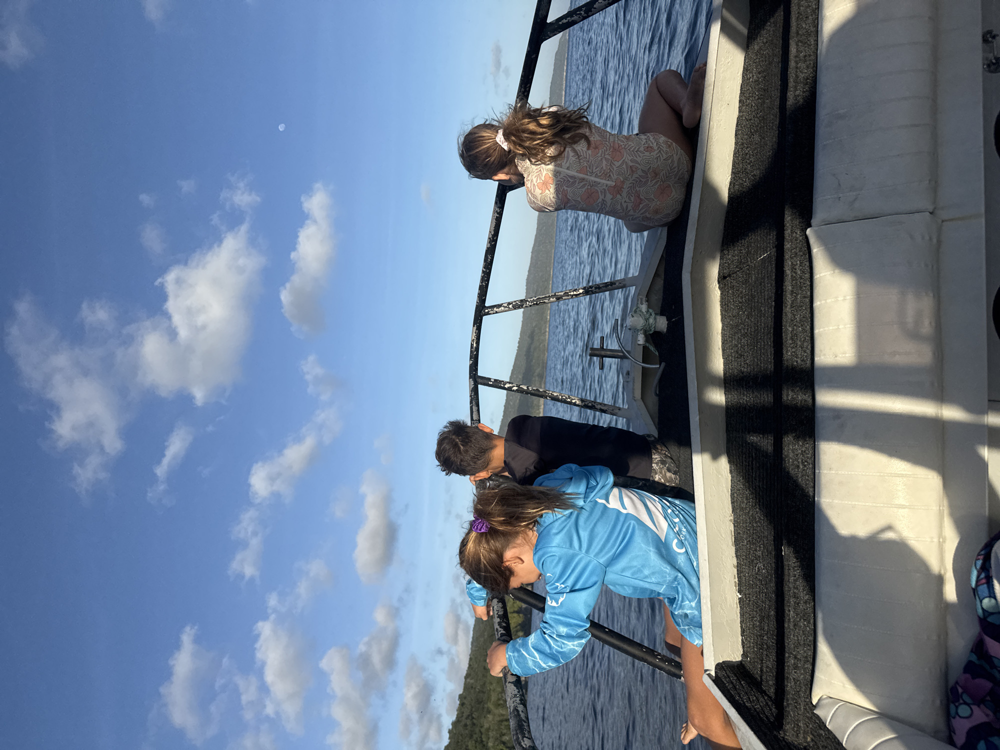 Three children, two girls and a boy, are on a boat on a body of water. The boat is moving, as indicated by the wake. The children are facing away from the camera. The girl on the left is wearing a blue long-sleeved shirt. The boy in the middle is wearing a black t-shirt and camouflage shorts. The girl on the right is wearing a swimsuit with a floral pattern. The background is a body of water with a tree-covered hill in the distance. The sky is blue with white clouds. The location is Christmas Island, Christmas Island.