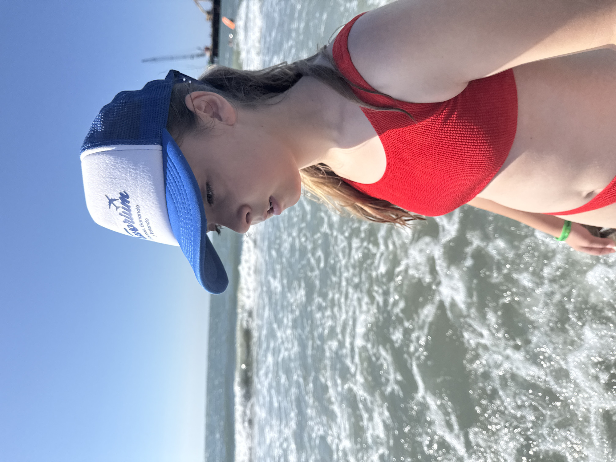 A young person wearing a red bikini top and a blue and white trucker hat is at the beach, partially submerged in the ocean. The water is choppy and reflecting the bright sunlight, suggesting a clear, sunny day, likely in the afternoon given the angle of the sun. The person is looking down and to the side, with their hair appearing damp and windswept. The hat has a logo with text on it. The visible text on the hat reads "Goribe" and "Ganando". The background shows a clear blue sky and the ocean stretching out to the horizon, with some distant structures that might be piers or boats. The overall scene evokes a sense of leisure and enjoyment of a beach day.