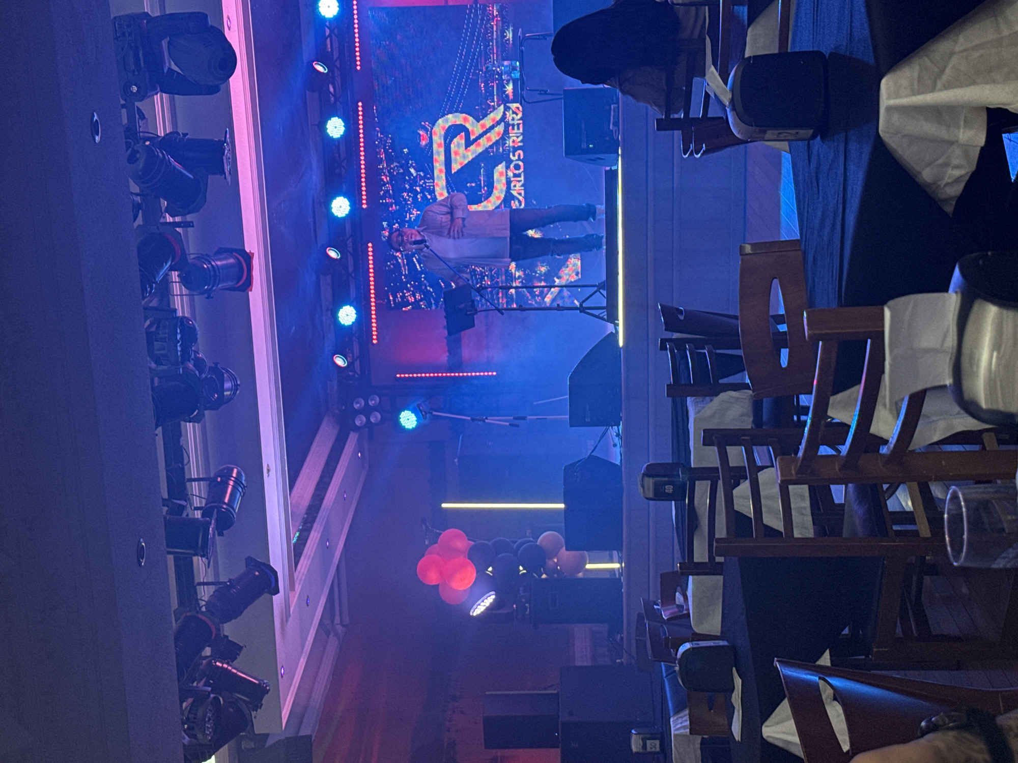The image depicts a performance taking place on a stage in a dimly lit venue, likely a restaurant or event hall, in Torrejón de Ardoz, Spain. The stage is adorned with a large LED screen displaying a logo with the letters "CR" and the name "CARLOS RIERA." A male performer, wearing glasses, a light-colored shirt, and jeans, stands at a microphone stand on the stage, actively speaking or singing. Stage lighting, primarily in blue and pink hues, illuminates the performance area. Several speakers and lighting fixtures are visible on and around the stage. In the foreground, tables with white tablecloths and chairs are set up, suggesting an audience seating area. A few patrons are partially visible in the seating area, with one individual in the lower right corner facing away from the camera. Red and black balloons are also present on the stage to the left of the performer. The overall atmosphere suggests a live entertainment event.