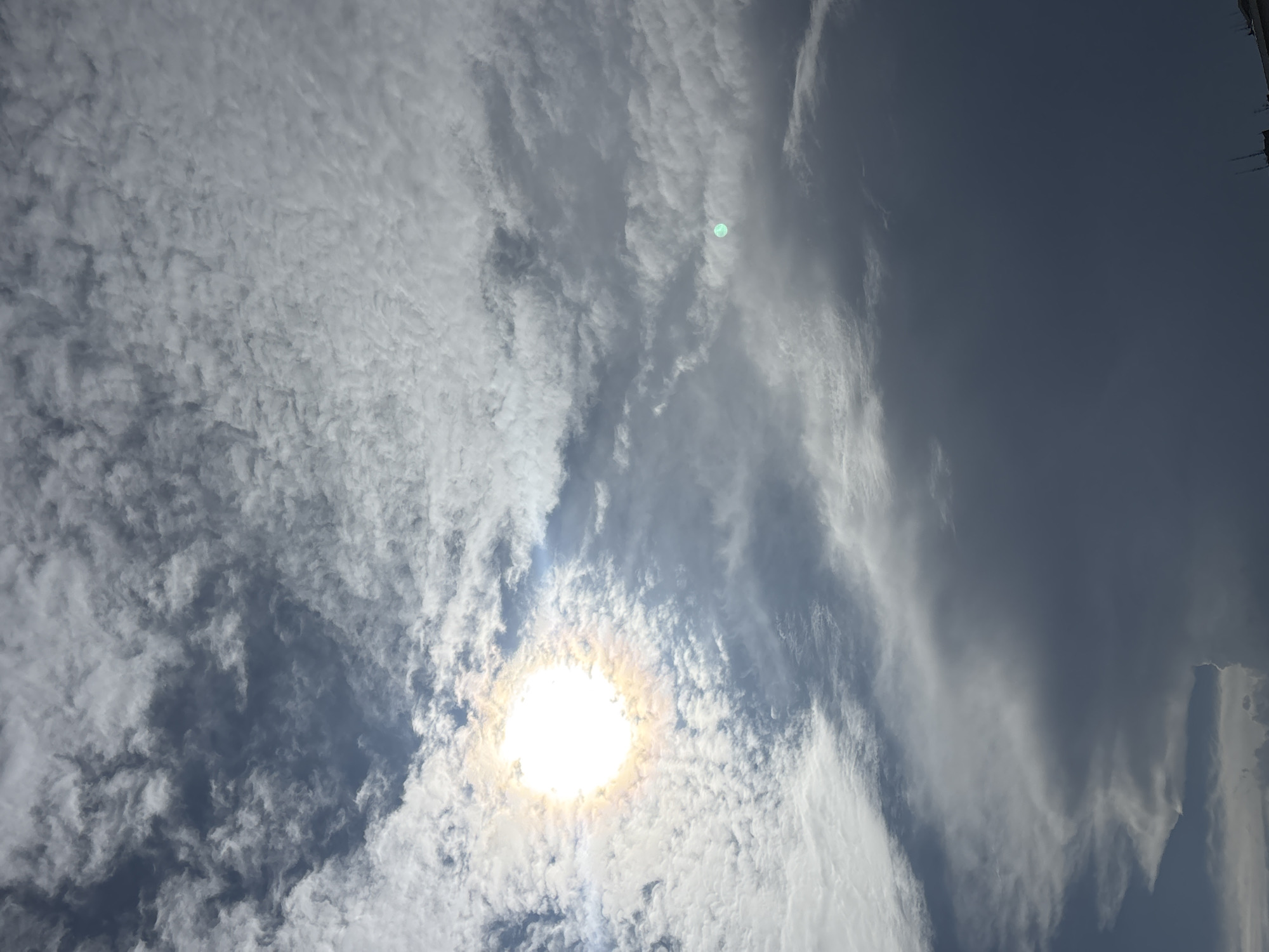 The image shows a view of the sky in Belgrade, Serbia. The sun is visible, partially obscured by a layer of altocumulus clouds. There are also cirrus clouds present on the right side of the image, indicating a change in the weather.
