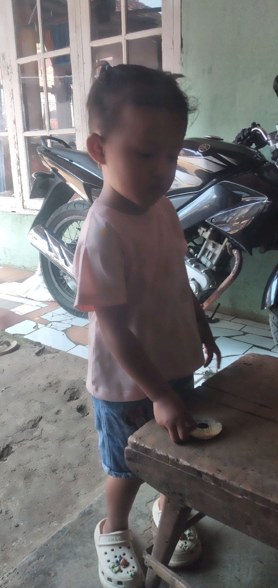 A young child, likely female, stands next to a wooden table and a motorcycle in an outdoor, possibly urban, setting. The child is wearing a light pink t-shirt and blue denim shorts. The child's hair is pulled back in a small ponytail. The child's right hand is resting on the table, near a small, round object. The child is wearing white clogs decorated with colorful embellishments. In the background, a black motorcycle is visible, with its wheel and exhaust pipe clearly distinguished. A white-framed window with multiple panes is also present. The ground is a mixture of dirt and tiled flooring. The scene appears to be located in Rahong, Indonesia.