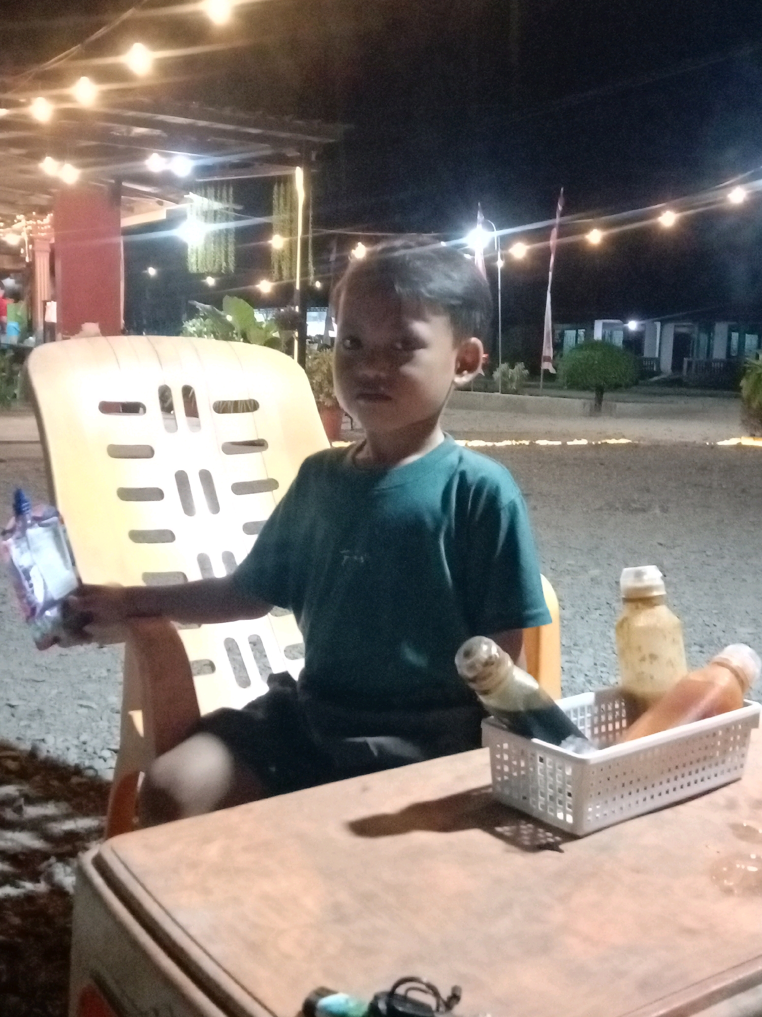 An Indonesian boy is sitting on a yellow plastic chair outdoors at night. He is wearing a dark green t-shirt and black shorts. He is holding a small, clear plastic packet in his left hand. In front of him, on a wooden table, is a white basket containing two bottles, one appears to be soy sauce and the other an orange-colored liquid. The background shows a dimly lit outdoor setting with string lights overhead. The location is identified as Utama Jaya Mataram, Indonesia.
