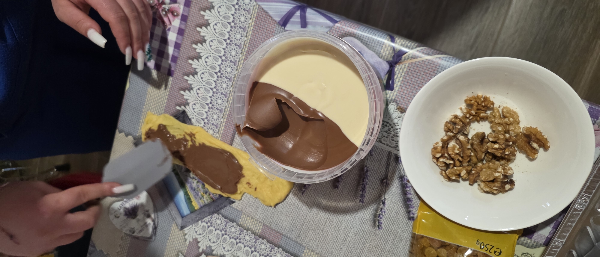 The image shows a partially assembled dessert on a patterned tablecloth, likely indoors. A person's hands are visible; one is holding a spatula and spreading chocolate-hazelnut spread onto a yellow pancake or crepe. The spread is from a large, clear plastic tub containing two distinct layers: one is brown chocolate-hazelnut, and the other is a light beige or vanilla spread. To the right of the tub is a white bowl filled with shelled walnuts. Below the bowl, a yellow package labeled "e250g" is visible. The scene is casual, suggesting food preparation or consumption. It is impossible to determine the location in Novachene, Bulgaria, or if it is even located there.