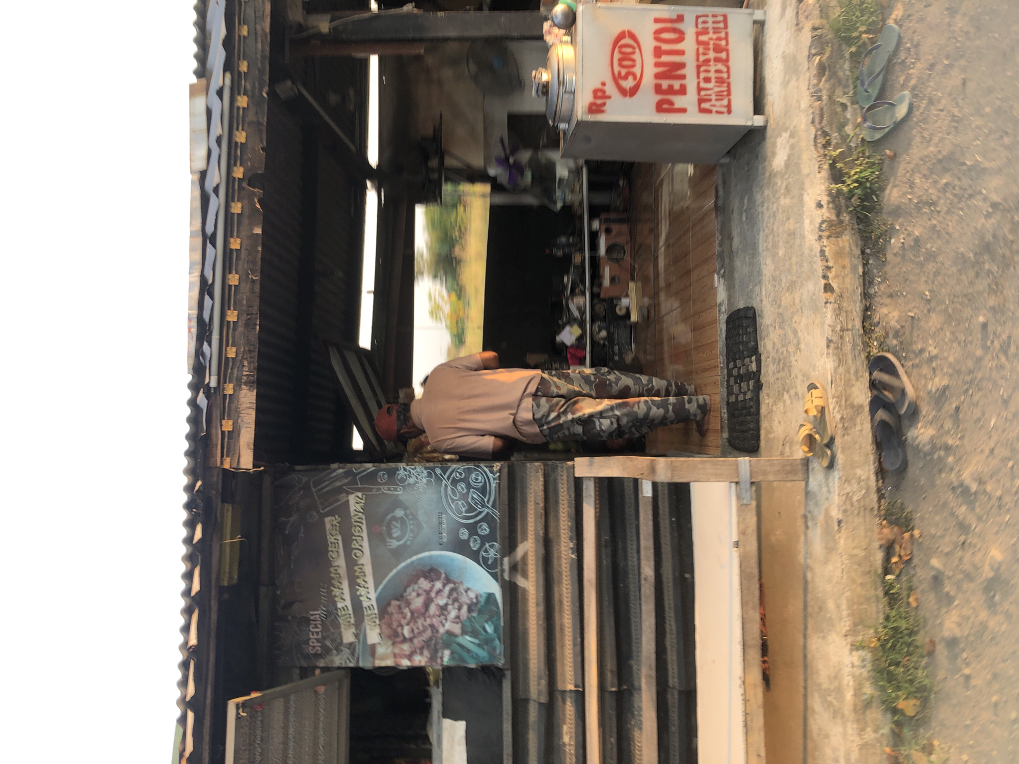 The image depicts a street-level view of a modest food stall located in Keboguyang, Indonesia. The stall features a corrugated metal roof with some exposed supports. To the left, a dark-colored sign displays "SPECIAL MENU" along with "MIE AYAM CEKER" and "MIE AYAM ORIGINAL" in white lettering, accompanied by an image of a bowl of noodles. A person wearing a light brown t-shirt and camouflage pants is standing in front of the stall, facing away from the camera and towards the interior. The interior of the stall is dimly lit and appears to house cooking equipment, including a large metal pot with a lid and a fan. To the right of the entrance, a white cooler box has red text that reads "Rp. 500 PENTOL". The ground in front of the stall is concrete, with some vegetation growing in cracks. Several pairs of flip-flops are visible on the ground, suggesting a casual, open-air establishment. The overall scene conveys a simple, functional food service operation in an outdoor setting.