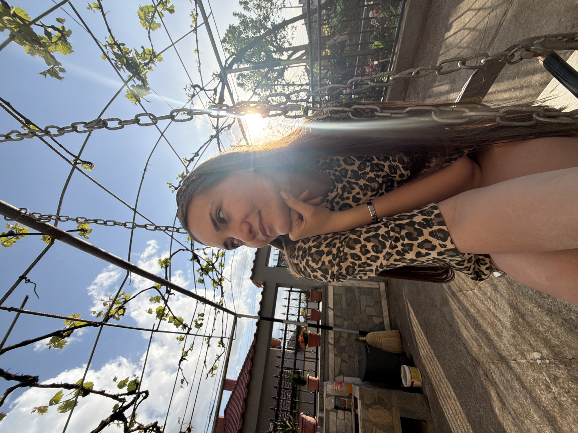 The image shows a woman with long brown hair wearing a leopard print top. She is resting her chin on her hand and appears to be posing for the picture. The background includes a metal chain structure, possibly a pergola or a trellis, with green vines and leaves. The sky is visible with some clouds, and sunlight is shining through the structure. In the distance, a building with a red roof can be seen, along with a stone wall, potted plants, and a broom. Based on the information provided, the location is Buhovo, Bulgaria.