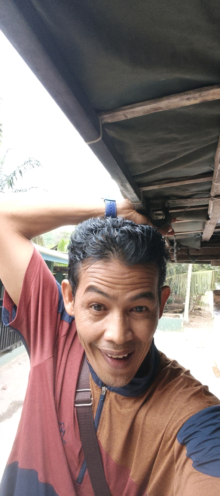 A middle-aged Indonesian man with dark, curly hair is taking a selfie. He is wearing a maroon and navy blue t-shirt with a brown zippered vest over it, and a brown strap across his chest. He has a blue watch on his left wrist. The man is smiling broadly, with his arm raised behind his head. He is outdoors, under a wooden and canvas structure, possibly a shelter or a vehicle. In the background, there is foliage and a hint of buildings, suggesting a semi-rural or suburban setting. The lighting is bright and natural. The image appears to have been taken in Baru, Indonesia.