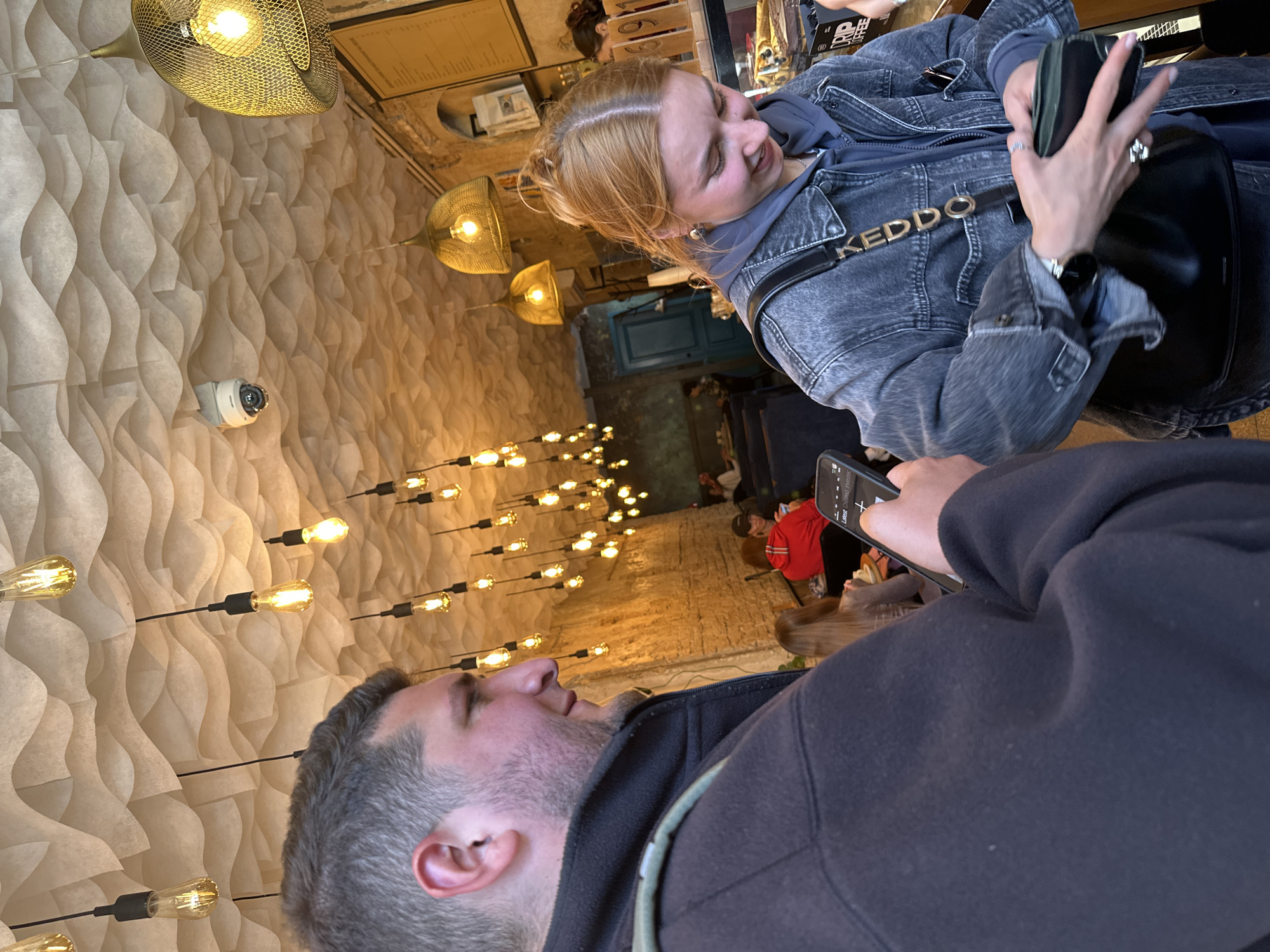 The image shows two people in what appears to be a cafe or restaurant. A man with graying hair is in the foreground, partially obscured by his own shoulder. He is looking upwards, possibly at the ceiling. The second person is a fair-skinned woman with reddish-blonde hair, wearing a denim jacket and a black bag with the word "KEDDO" on the strap. She is holding a black wallet or phone and looking down, possibly at the man or the phone in his hand.
In the background, there are other patrons seated at tables. The decor includes an undulating white textured wall and multiple light fixtures with exposed bulbs. From the style of decoration, it may be similar to modern cafes found in the city of Hrodna, Belarus. The people appear to be engaged in a casual conversation or interaction. A security camera is on the textured wall.