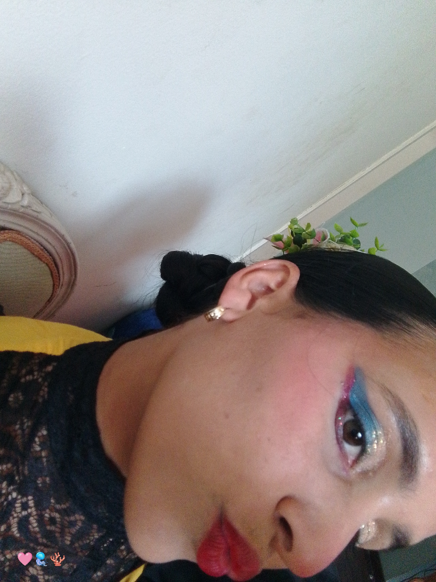 A close-up, indoor shot shows a person with dark hair pulled into a bun. They have on bold makeup: red lipstick, pink blush, and blue and silver eyeshadow. The environment appears to be a room with white walls and a section of decorative trim at the top. A small cluster of artificial pink flowers and green leaves is tucked into their hair. The exact location in Pedro González, Venezuela, is not discernible from the image.