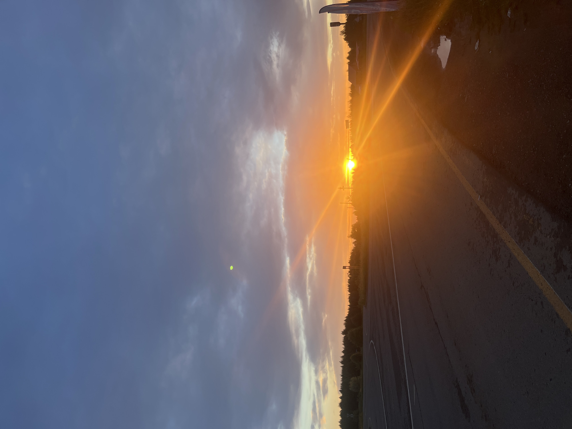 The image depicts a sunset over a paved road in Gol'yany, Russia. The sky is filled with dramatic clouds, ranging from dark gray to vibrant orange as the sun dips below the horizon. The sun's rays are casting a warm glow across the scene, illuminating the road and the surrounding trees. The road appears to be an intersection or a parking lot, with visible lane markings and a slight curve. There are no people visible in the image. A few structures, possibly buildings or utility poles, can be seen in the distance. The overall impression is one of a serene and picturesque natural landscape.