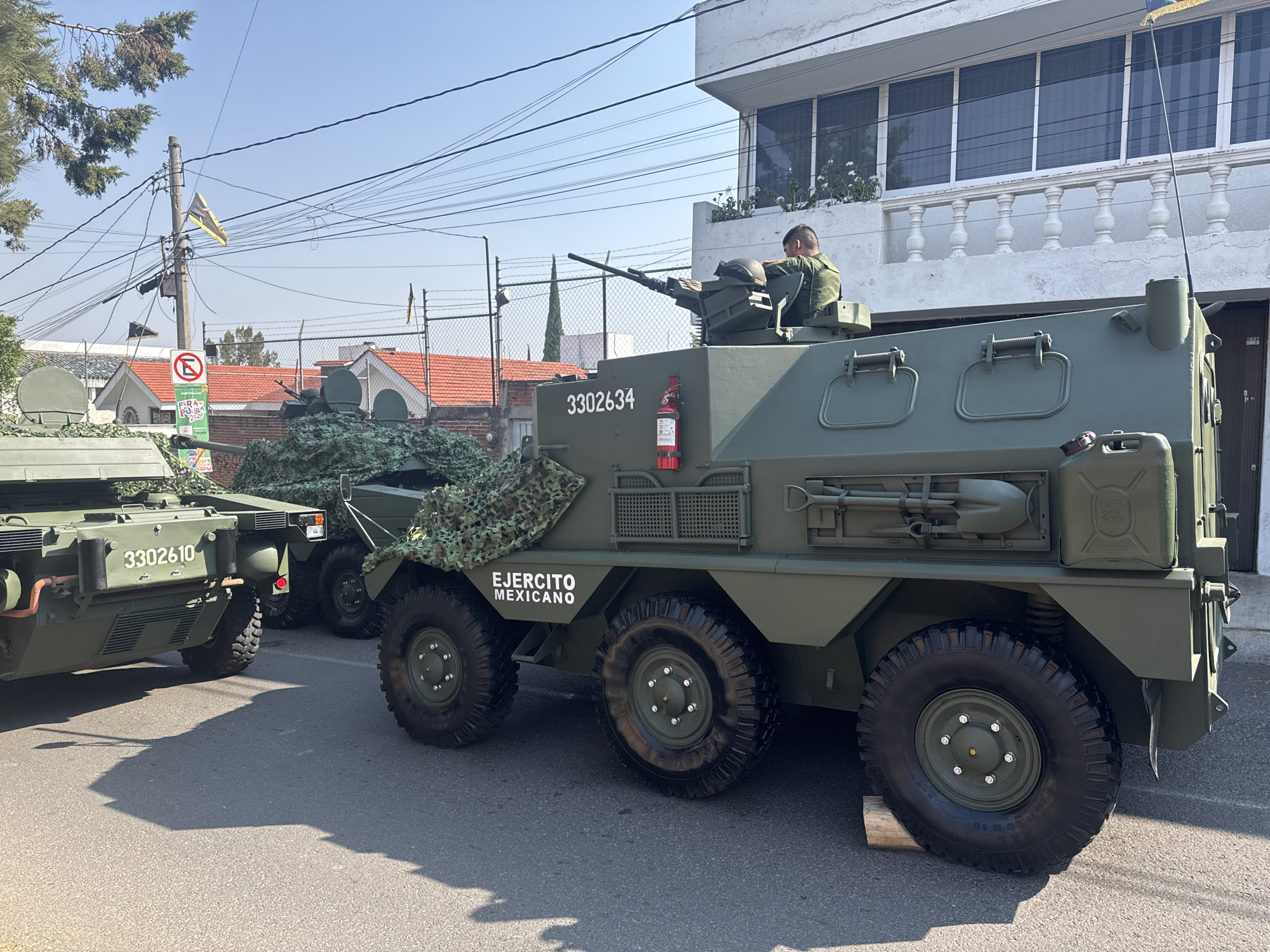 The image shows a line of olive-green military vehicles, likely AML Panhard or similar armored cars, parked on a city street. The vehicle closest to the camera is marked with the number "3302610". The second vehicle is numbered "3302634" and has a soldier in a military uniform manning a weapon turret on top. The words "EJERCITO MEXICANO" are visible on the side of the second vehicle. The vehicles behind the first two are partially obscured.
The background features residential buildings. Power lines and a traffic sign are also visible. The presence of "EJERCITO MEXICANO", along with the signs in Spanish, indicates the location is in Mexico. The presence of the traffic sign with "Feria de Puebla 2023" suggests that the vehicles are located in Puebla, Mexico. The lighting indicates it is daytime.