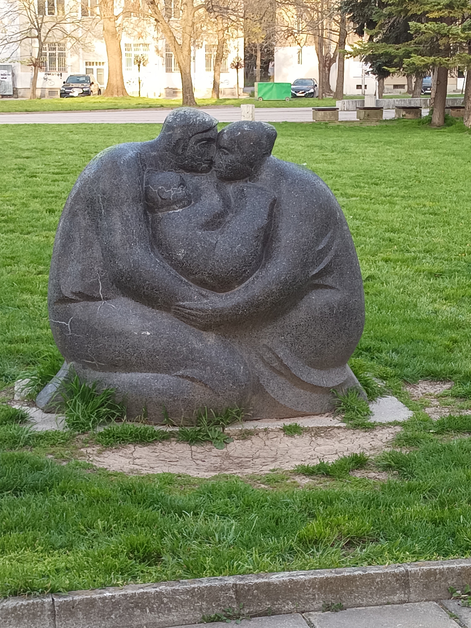 A sculpture depicting a family group is centrally featured in this vertical shot, set within a park-like environment. The sculpture, made of dark gray stone, portrays two adults embracing tenderly, with a small child nestled between them. The figures are stylized and rounded, conveying a sense of warmth, unity, and protection. Visible cracks on the surface, particularly on the left figure's hip, indicate age or weathering of the stone.
The sculpture rests on an irregular concrete base, surrounded by a mix of vibrant green grass and patches of dry, cracked earth. In the foreground, a concrete curb is partially visible at the bottom edge.
Beyond the immediate grassy area, the background reveals more of an urban park setting. Bare deciduous trees, suggesting early spring or late autumn, stand alongside some evergreen conifers. Further back, buildings with multiple windows and balconies line what appears to be a street or open area. A black car is visible in the mid-ground, parked near one of the buildings, and another car is partially seen behind trees. A green dumpster or utility box is also present on the left side of the background.
The lighting suggests late afternoon or early morning, casting soft light on the scene. No specific weather conditions are discernible, other than it being daylight. There is no visible text in the image. The setting is consistent with a public space in Targovishte, Bulgaria.