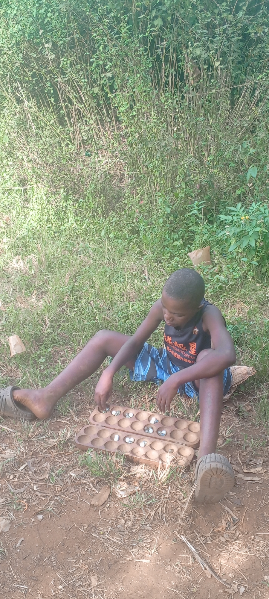 A young person sits on the ground in Ngalula, Democratic Republic of the Congo, engaged in playing a game. The game board, a wooden mancala board with round, light-colored stones, sits between the person's outstretched legs. The individual, dressed in a dark tank top and patterned shorts, appears focused on the game. The setting is a grassy area with dense foliage in the background. One of their feet is clad in a sandal. The ground is a mix of grass and dirt. A discarded paper bag is visible near the foliage.