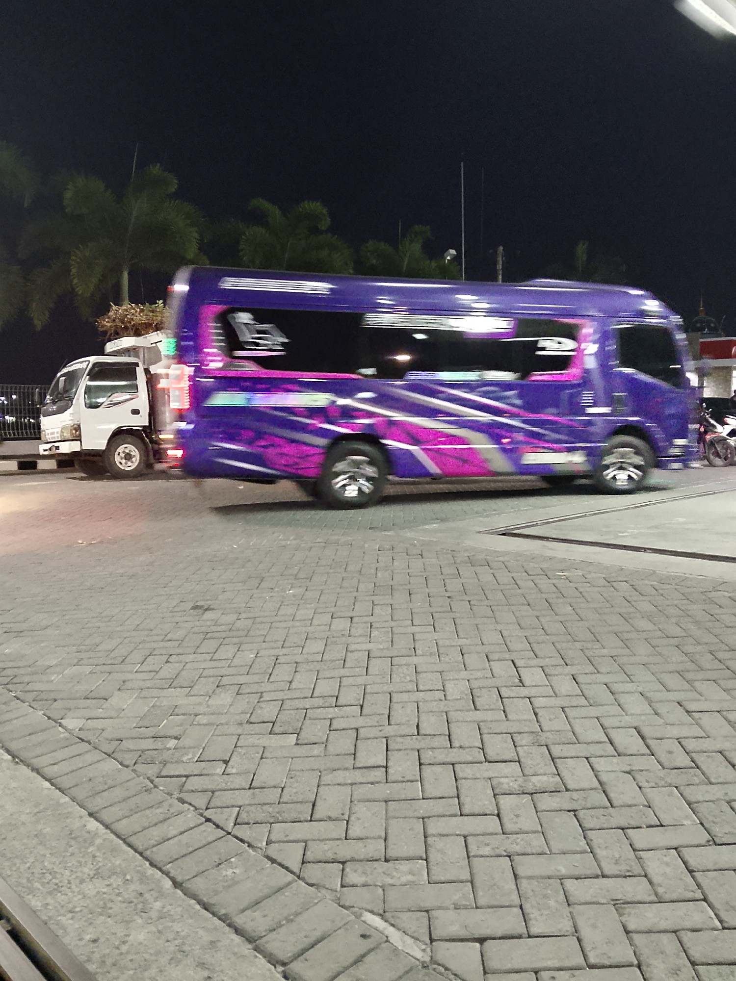 A purple bus with pink and silver graphic designs on its side is parked next to a white truck. The bus is a passenger van with tinted windows, one of which shows a silhouette of seats inside. The truck is a small cargo vehicle with its headlights and front grill visible. The vehicles are parked on a paved surface made of interlocking rectangular bricks, laid in a herringbone pattern. In the background, palm trees are silhouetted against a dark night sky. A motorcycle is parked to the right of the bus. The scene appears to be in an outdoor urban setting at night, possibly a parking area or roadside in Kasiman, Indonesia, given the presence of palm trees and the typical brick paving.