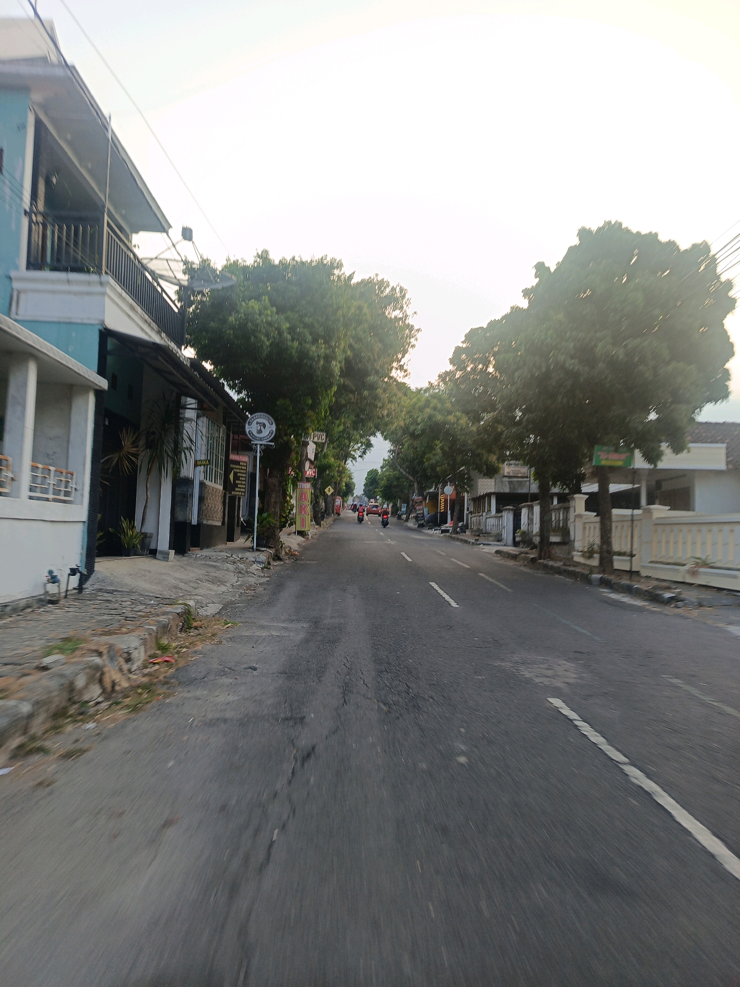 The image shows a street scene in Magetan, Indonesia, likely taken from a moving vehicle. The road is paved and has a white line marking the lane. On the left side of the road, there are buildings with balconies and satellite dishes visible. One building is painted turquoise and white. Further down the street, there are more buildings and trees lining the road. Several motorcycles are visible on the road, with some people riding them. The sky is bright and overcast, suggesting it might be early morning or late afternoon. The overall impression is a typical residential or commercial street in a town or city.