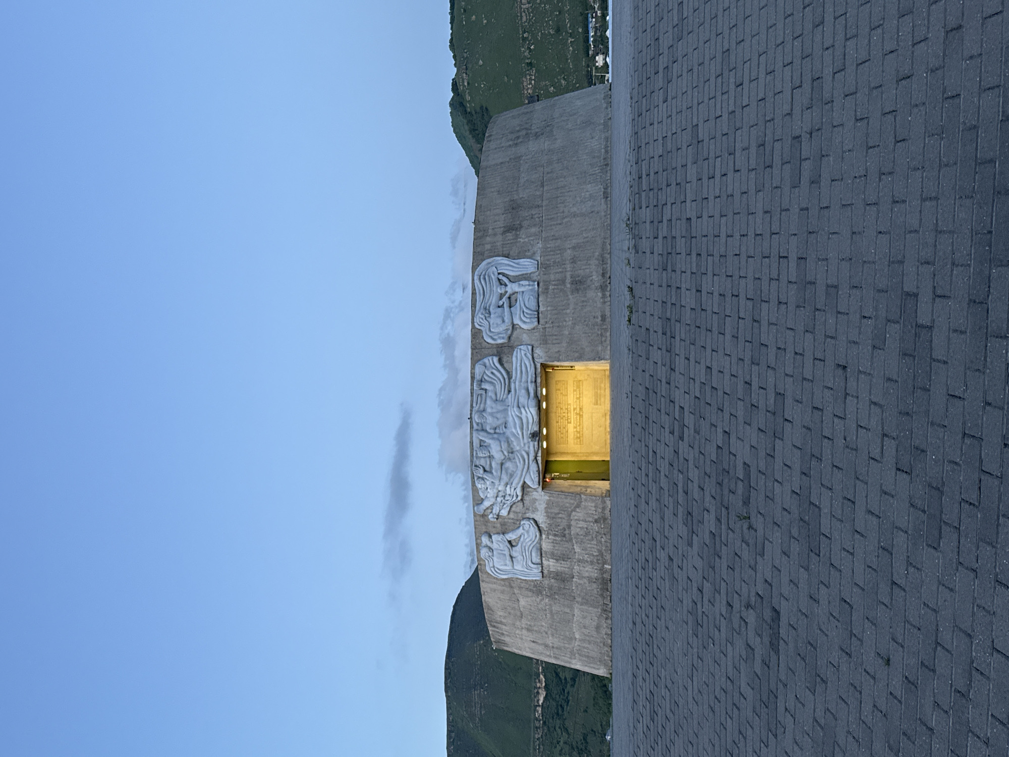 The image shows a concrete structure with bas-relief carvings located near a mountain range under a blue sky. The structure has an illuminated doorway. A paved area extends to the right of the structure. The text on the wall is the name of the City of Ordzhonikidzevsky in Russia in the Ossetian language.