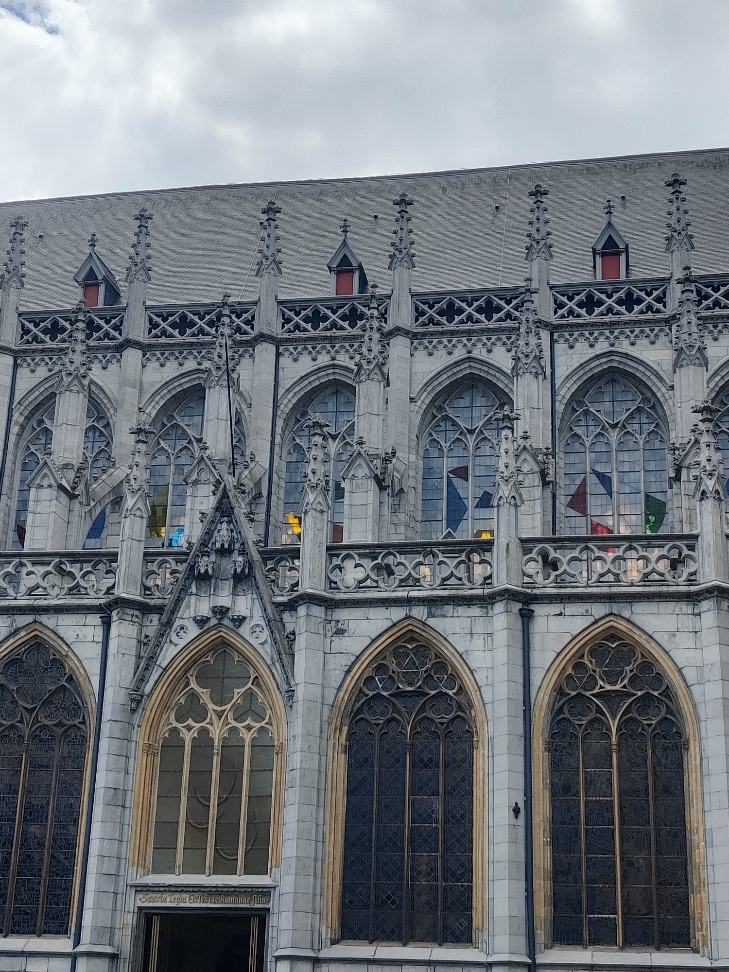The image shows the exterior of a large Gothic-style building, likely a church or cathedral, with intricate stonework and arched windows. The building is made of light-colored stone and features decorative spires and ornate window tracery. The windows themselves are large and appear to be made of stained glass, with some showing colored patterns and others filled with dark, presumably leaded, glass. There are also small dormer windows visible on the steeply pitched roof. The sky above is overcast and grey. A sign with text is visible above a doorway on the lower level. This building is the Saint Paul's Cathedral (Cathédrale Saint-Paul) in Liège, Belgium.