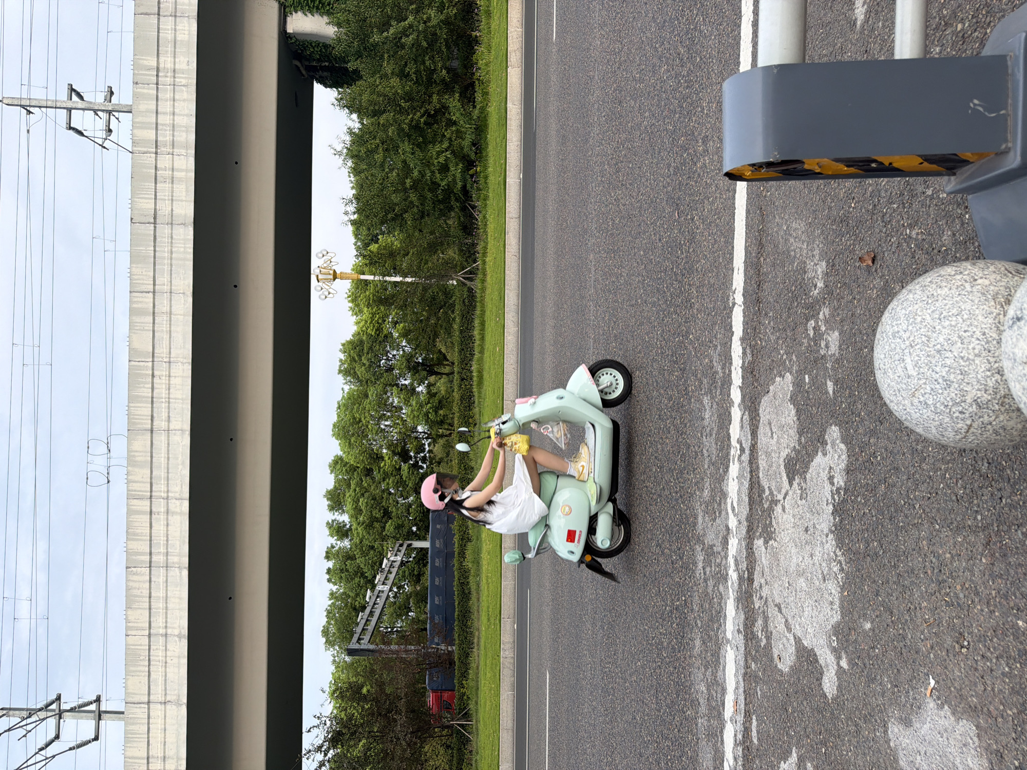 A young girl wearing a white dress and a pink helmet rides a mint green electric scooter on a paved road in Jiangjin, China. The scooter has a yellow bag hanging from the handlebars and a small flag on its side. The road is lined with green grass and trees, with an elevated highway and power lines visible in the background. The sky is overcast.