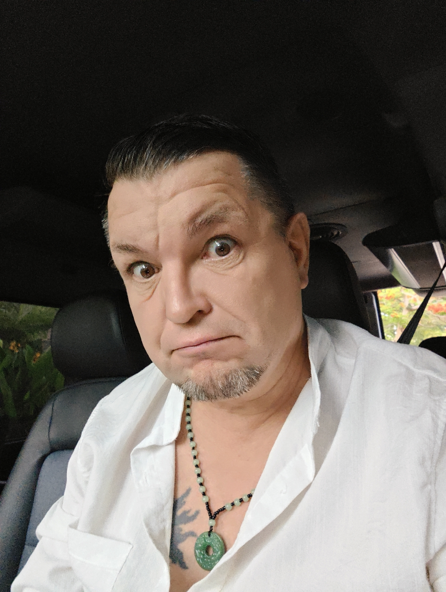 The image is a close-up, selfie-style photo of a man with short, dark hair, styled back from his forehead. He has light brown eyes, raised eyebrows, and his mouth is downturned, giving him a surprised or worried expression. He is wearing a loose-fitting, white collared shirt, open at the neck, revealing a tattoo on his chest and a necklace with green jade pendant. He appears to be in a vehicle, as indicated by the dark interior of what looks like a car or SUV with black leather seats visible behind him, and a rearview mirror mounted above his head. Through the windows, foliage and trees are visible, suggesting an outdoor location. The background is blurred, but appears to be lush and green, possibly in a tropical or subtropical environment. There is no specific indication of the location being in Bac Nha Trang Ward, Vietnam, within the image itself; this information is provided externally.