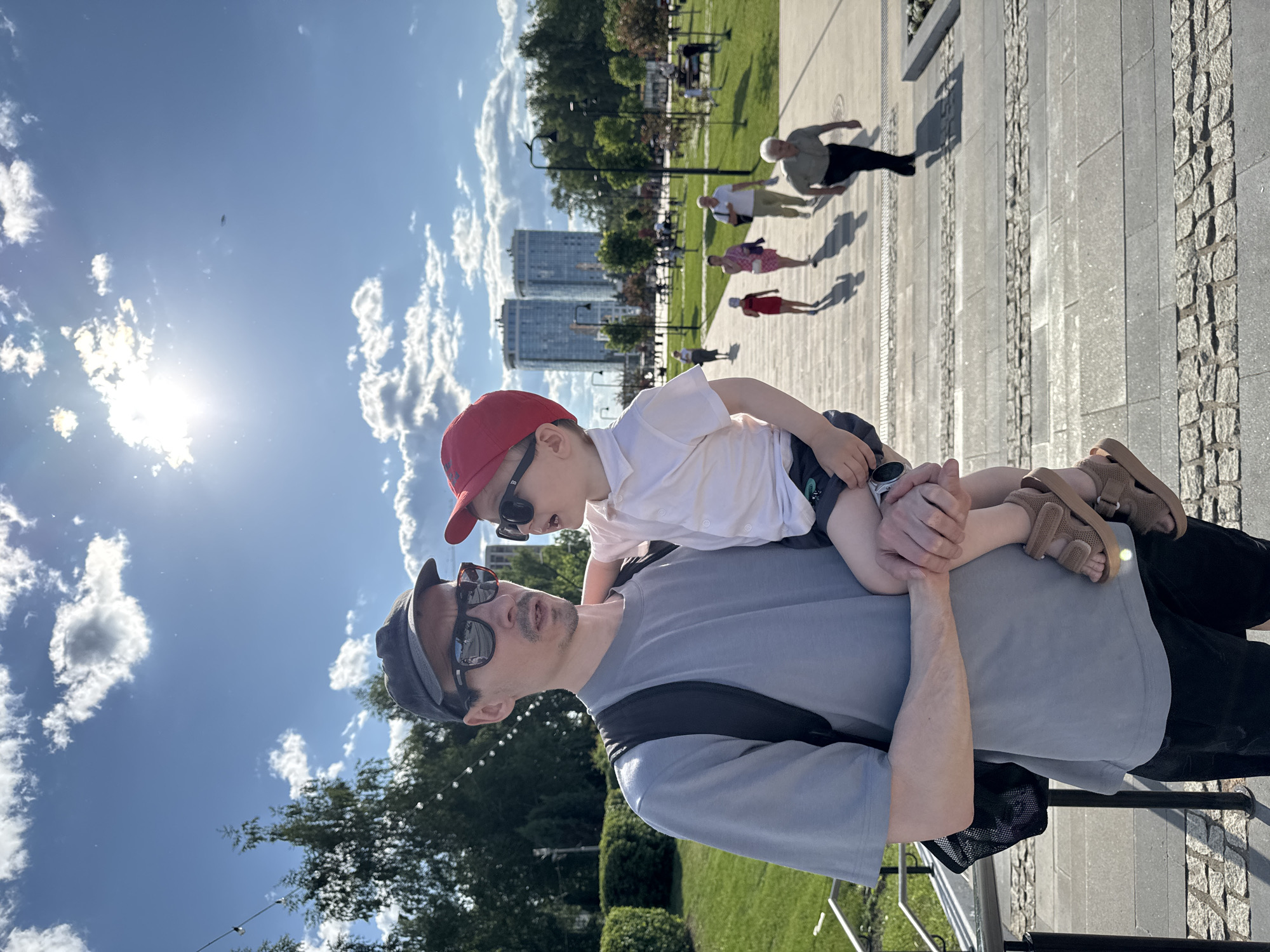 This image depicts a man holding a child in an outdoor urban park or plaza during daylight hours on a sunny day.
The primary subject is an adult male, appearing to be in his late 20s to 30s, with short dark hair and a mustache/goatee. He wears a grey baseball cap, dark sunglasses with a visible reddish frame, a light grey short-sleeved t-shirt, dark shorts, and a dark backpack. He holds a young child in his left arm. His head is slightly turned towards the child.
The child is a toddler, estimated to be between two and four years old. The child wears a red baseball cap, dark sunglasses, a white short-sleeved collared shirt, dark shorts, and brown sandals. The child is positioned facing the man, looking upwards at him with an open mouth, suggesting interaction or speech. The child's left hand grasps a small silver-colored object.
The setting is a well-maintained park featuring wide paved pathways and steps constructed from light grey and darker grey stone or cobblestone. A metal railing is visible in the lower-left corner. Green lawns, manicured bushes, and various deciduous and coniferous trees with summer foliage populate the background. String lights are suspended between trees on the left side of the frame. In the mid-ground, several individuals, including adults and possibly another child, are seen walking along the paths, casting distinct shadows due to the strong sunlight.
The sky is clear and blue with scattered white cumulus clouds. A bright sun flare is prominent in the upper right quadrant of the image. In the far distance, beyond the parkland, two tall, modern, glass-fronted high-rise buildings are discernible against the skyline.
Based on the distinctive architecture of the high-rise buildings in the distance and the style of the urban park, the location is identified as Izhevsk, Russia, consistent with modern developments and public spaces within the city, such as those near the central square or the "Izhevsk Plaza" area.