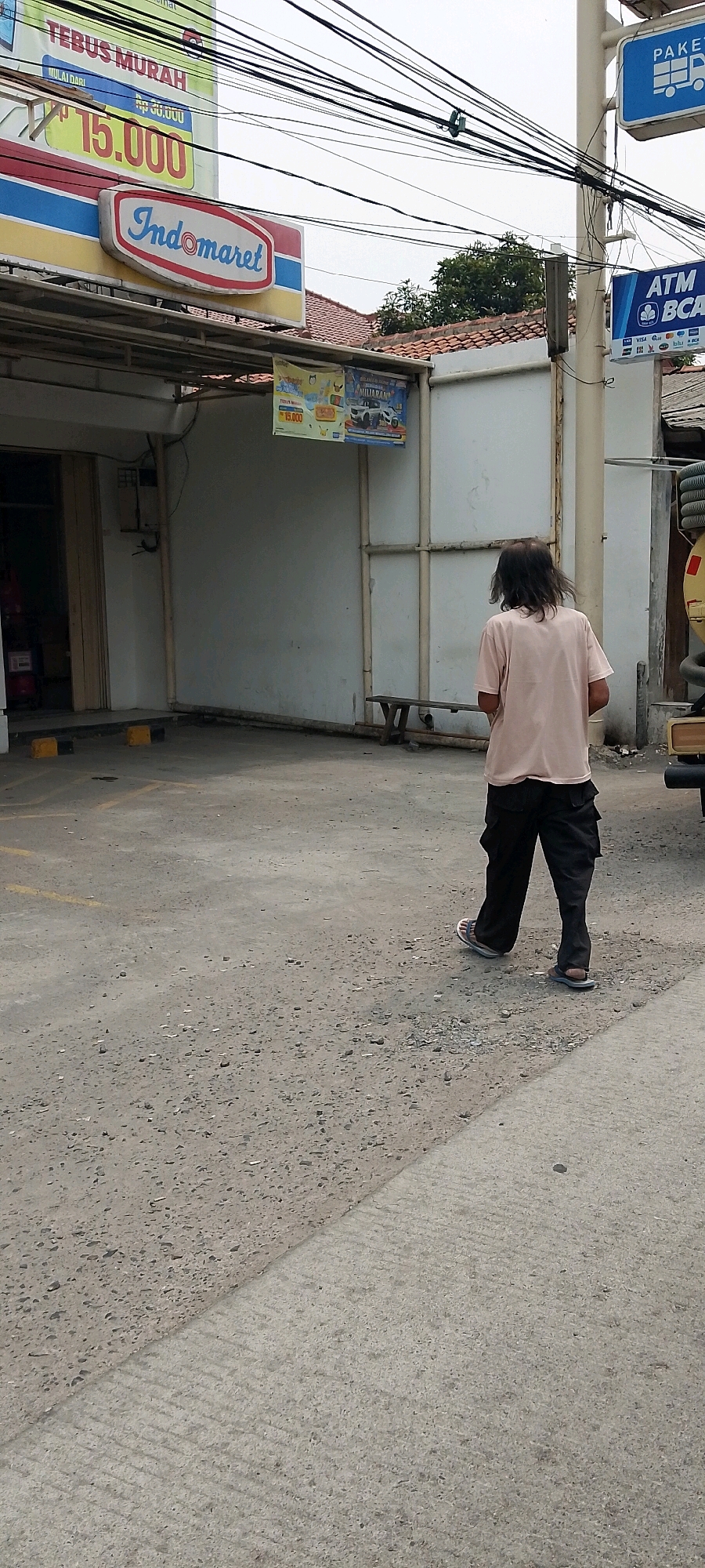 In the city of Depok, Indonesia, a man with long, unkempt dark hair, wearing a light pink t-shirt and dark cargo pants, walks away from the camera on a textured concrete ground. He is wearing light-colored sandals. To his left, a building facade is visible with signage indicating "Indomaret" and "TEBUS MURAH" with a price of "Rp 15.000". Further to the right, a white pole supports blue signs, one with a truck icon and "PAKE", and another with "ATM BCA". The environment appears to be an outdoor commercial area, possibly a parking lot or walkway in front of a retail establishment, with electrical wires strung overhead.