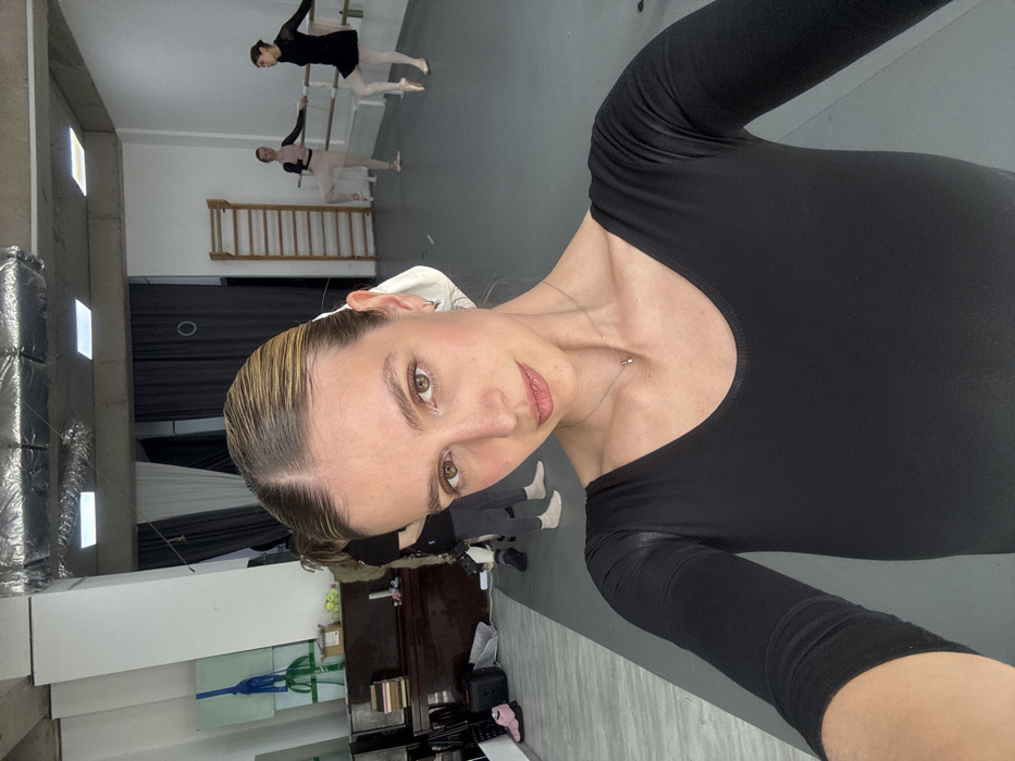 This image depicts an interior scene, identified as a dance studio, in the city of Unknown, Unknown. In the immediate foreground, a light-skinned female with light brown hair pulled back into a ponytail, wearing a black long-sleeved leotard and a silver necklace with a small pendant, is taking a selfie, looking directly at the camera.
In the background, to the left, a dark brown upright piano is visible with books or papers stacked on its top. A partially obscured individual, wearing dark trousers and a dark top, with bare feet, stands facing away from the camera near the piano. A pair of pink athletic shoes and a green, folded mat are on the floor in this area. To the right, two additional females, dressed in ballet attire, are actively practicing at a wooden ballet barre. The dancer on the left wears a light-colored leotard with a black chest portion and light-colored tights, holding an elevated leg. The dancer on the right wears a black leotard, a black skirt, and pointe shoes, also holding an elevated leg. A wooden wall bar system is mounted on the far right wall.
The environment consists of white walls, a grey floor, and a ceiling featuring exposed silver-colored ductwork, electrical conduits, and rectangular recessed lighting fixtures. Dark curtains are visible on the left side of the studio.
