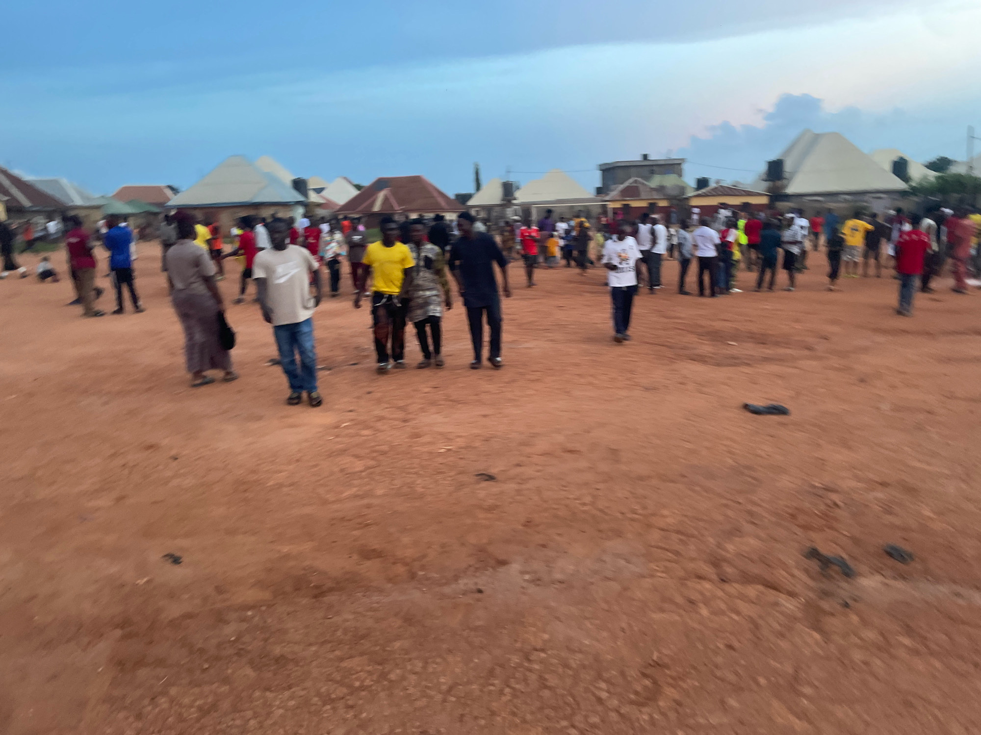 The image depicts an outdoor gathering of approximately 30-40 people in the city of Kurunduma, Nigeria. The ground is a reddish-brown, dusty earth. In the background, several buildings with sloped, light-colored roofs are visible. The sky is overcast with a pale blue hue and scattered clouds.
The people are standing and walking around, with no clear focal point or organized activity. They are dressed in casual clothing, with a variety of colors and styles. Some individuals are standing in small groups, while others appear to be walking individually. The lighting suggests it is either early morning or late afternoon. The overall impression is that of a community gathering or an informal event taking place in a semi-urban or rural setting.
There are several small, dark objects scattered on the ground, appearing as debris or refuse. One particular cluster of these objects is visible in the foreground. The image quality is somewhat blurry, making it difficult to discern fine details of the individuals or their expressions.