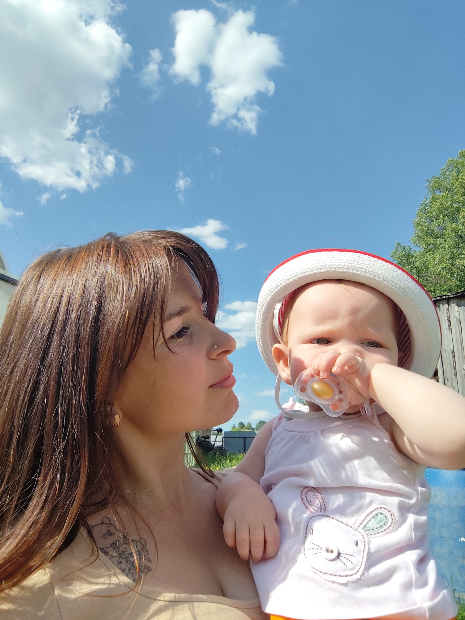A woman holds a baby outdoors on a sunny day, likely in Фоки, Russia. The woman has brown hair and a tattoo on her chest. The baby is wearing a red and white hat and has a pacifier in its mouth. The background includes a blue sky with white clouds, a wooden fence, and some greenery.