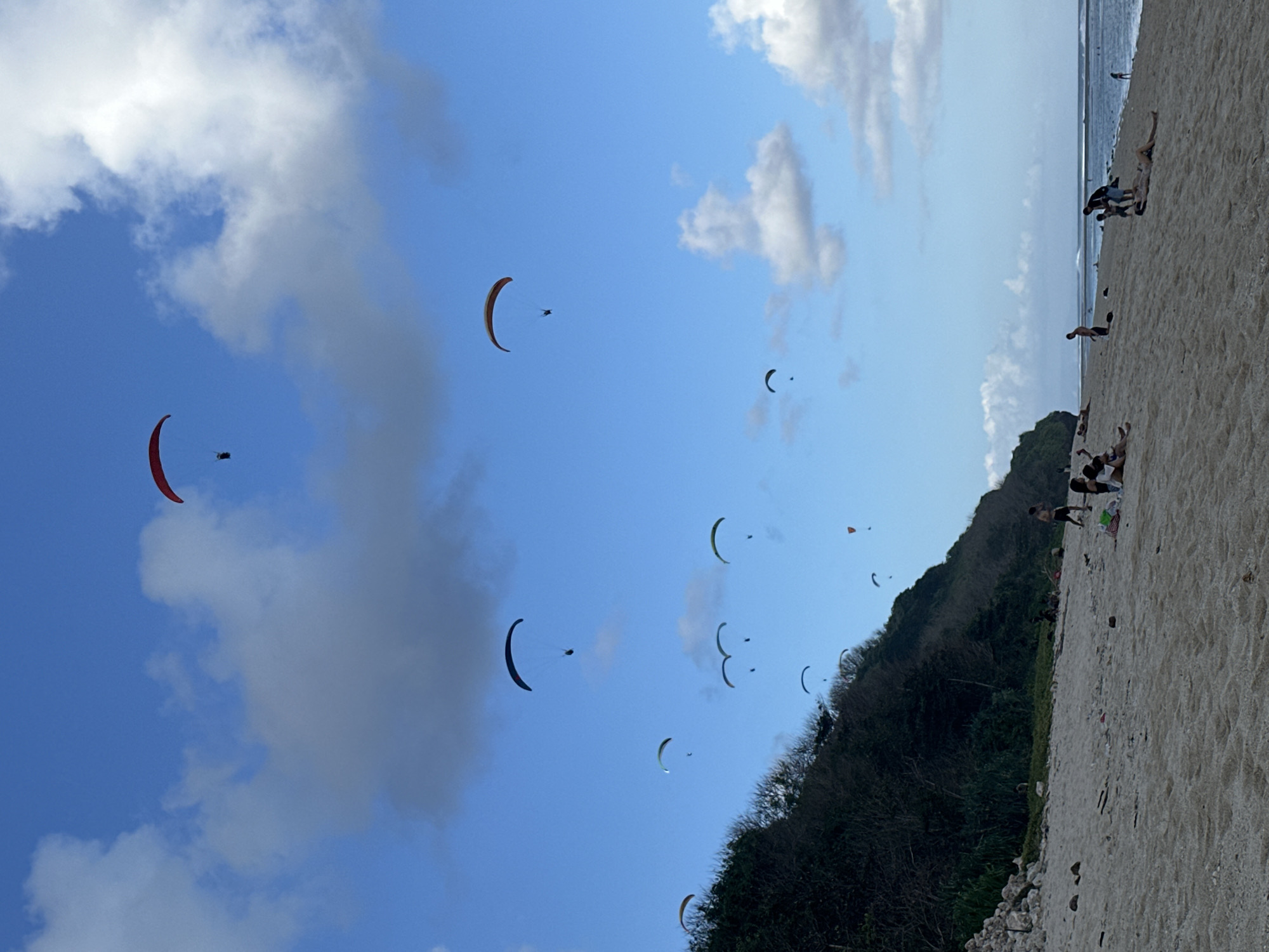 The image depicts a wide, sandy beach in Kuta Selatan, Indonesia, with the Indian Ocean in the background. The sky is a bright blue with scattered white clouds, and several paragliders are visible in the air, some with vibrant colored wings. On the beach, there are a few individuals and small groups of people, some sitting or lying down, while others are standing and walking along the shoreline. The environment includes a lush, green hillside on the left side of the image, meeting the beach. The overall scene suggests a recreational day at the beach with paragliding activities taking place.